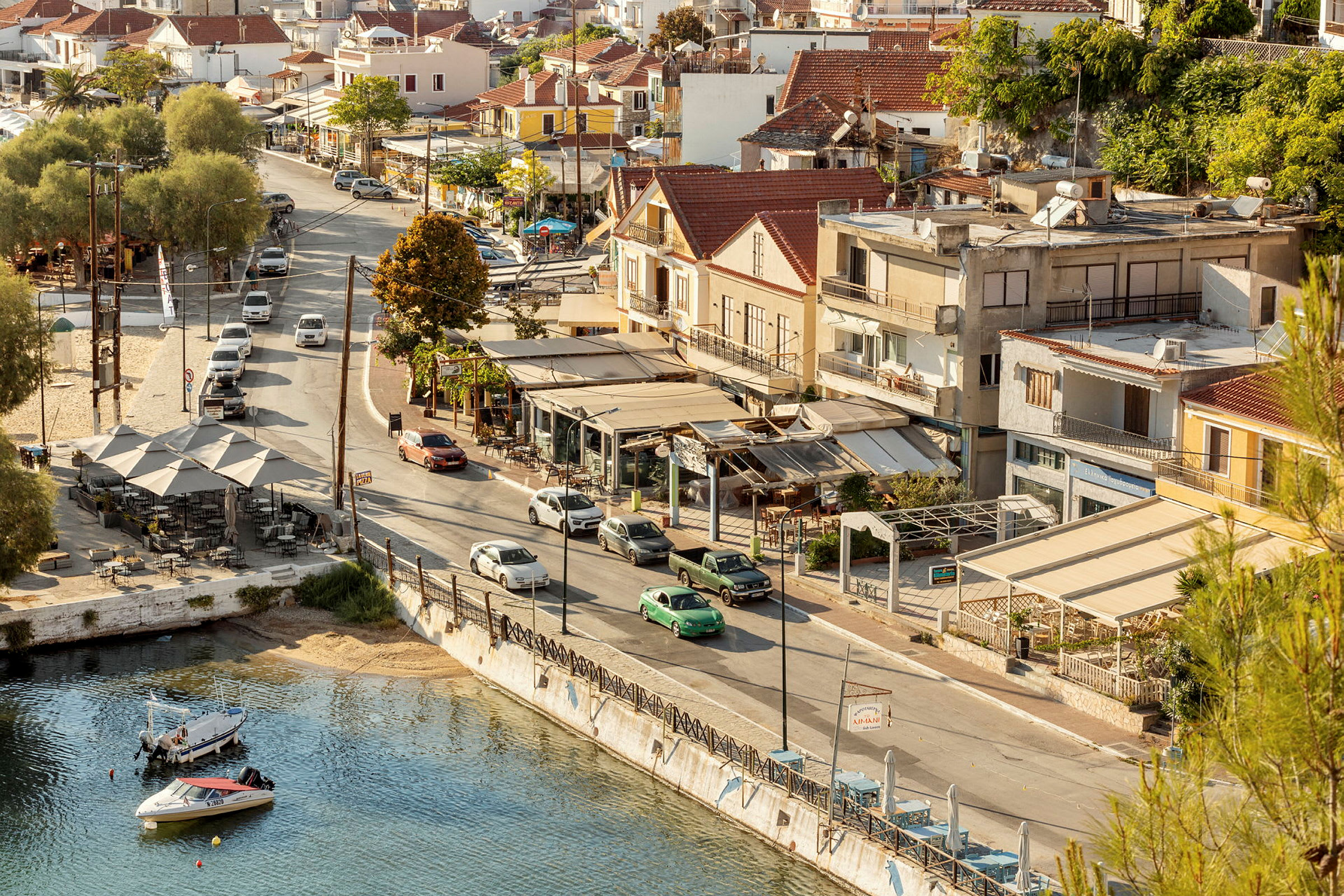 Vy över en stadsgata med bilar, hus, restauranger och vatten med små båtar. (AI-genererad text)
