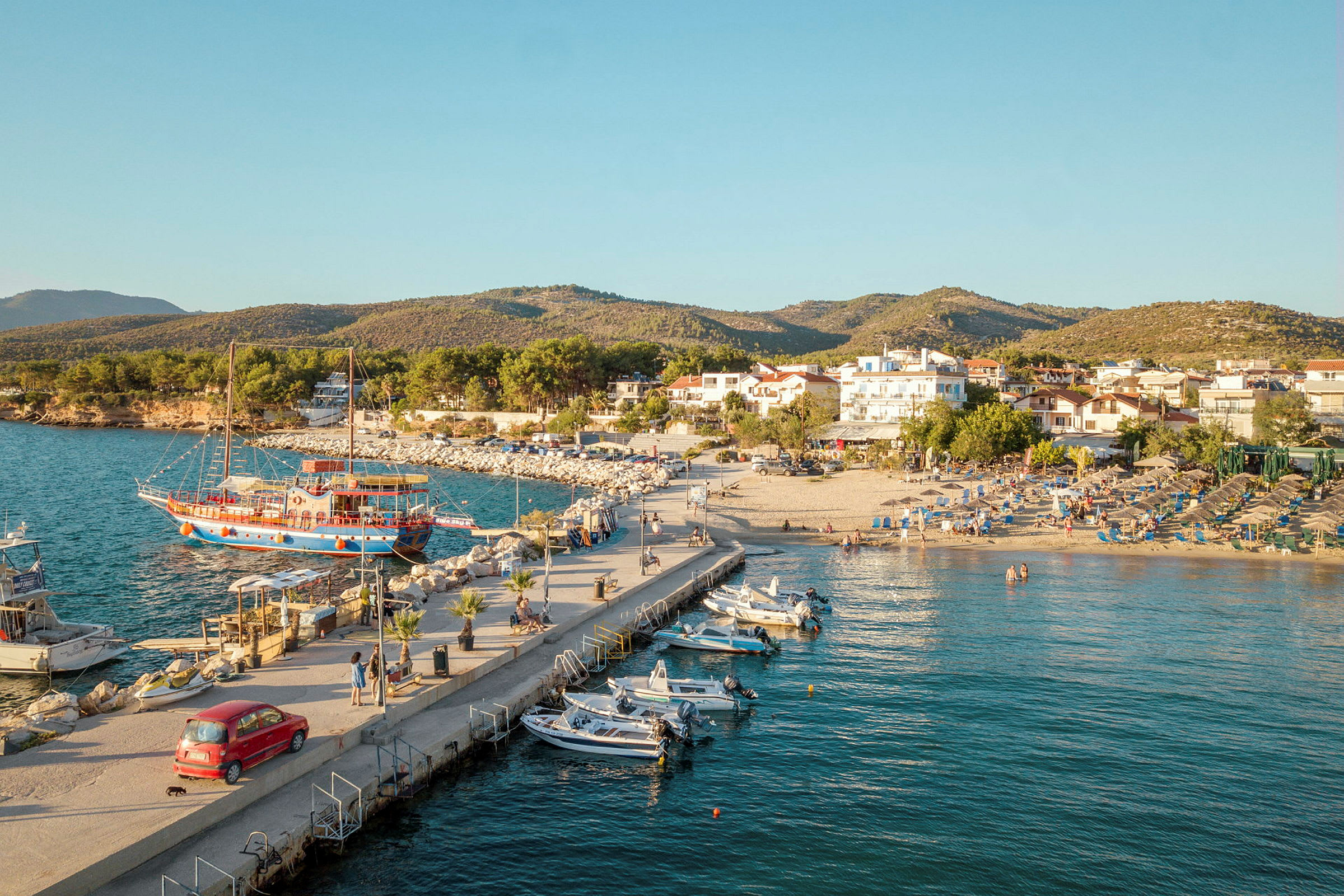 Vy över en hamn med båtar, en strand med solstolar och hus i bakgrunden. (AI-genererad text)
