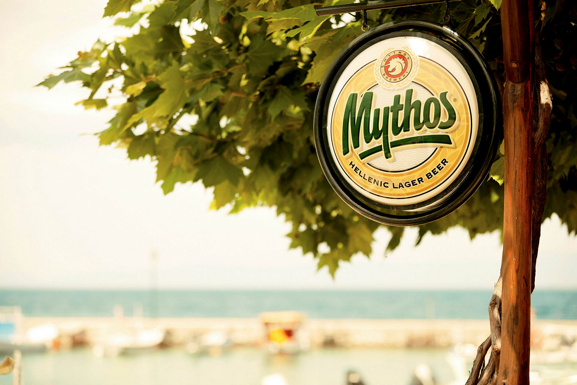 En hängande skylt med texten 'Mythos Hellenic Lager Beer' framför en suddig bakgrund av ett träd och en kustlinje. (AI-genererad text)