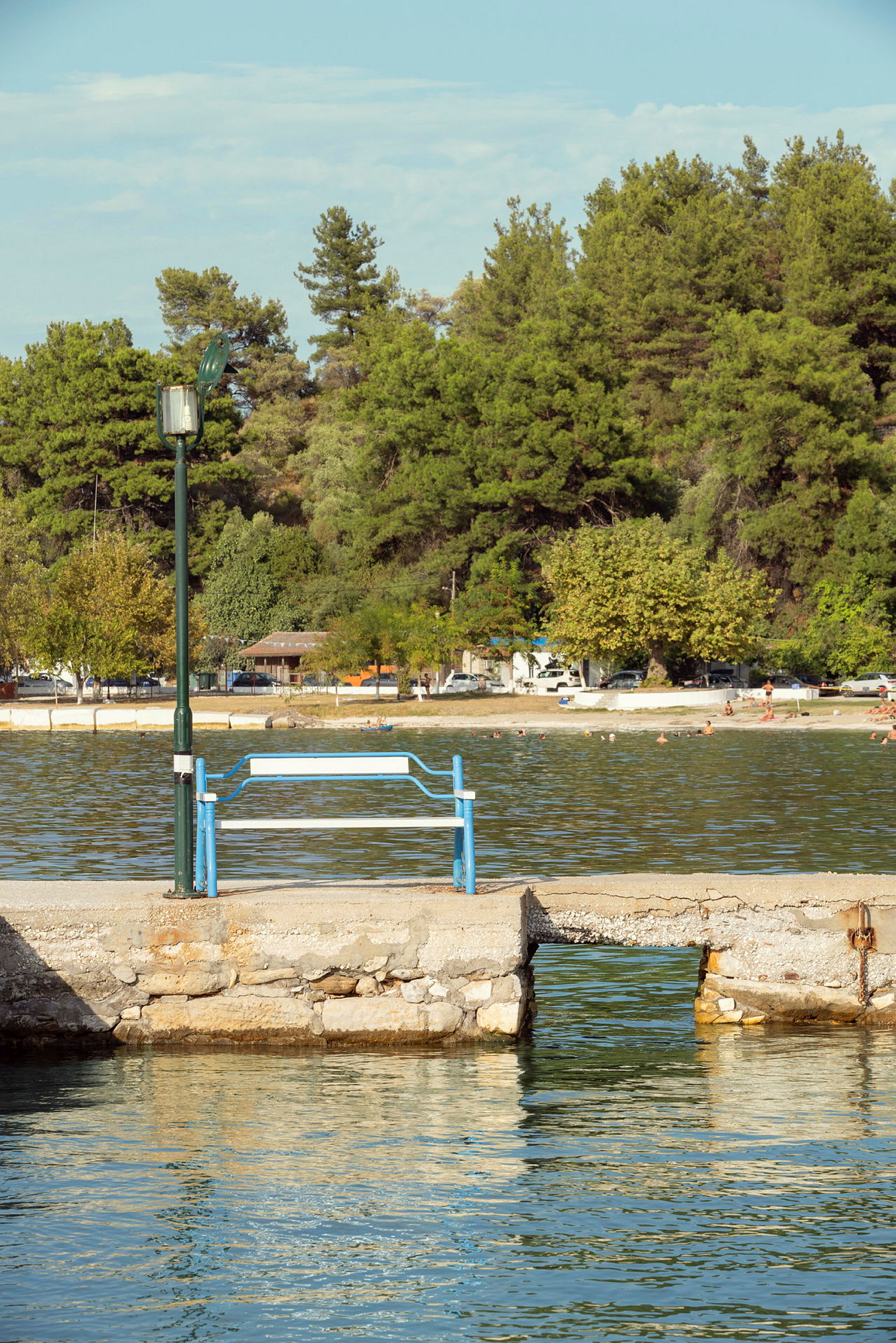 Brygga med en bänk och lyktstolpe vid vatten med skog och strand i bakgrunden. (AI-genererad text)