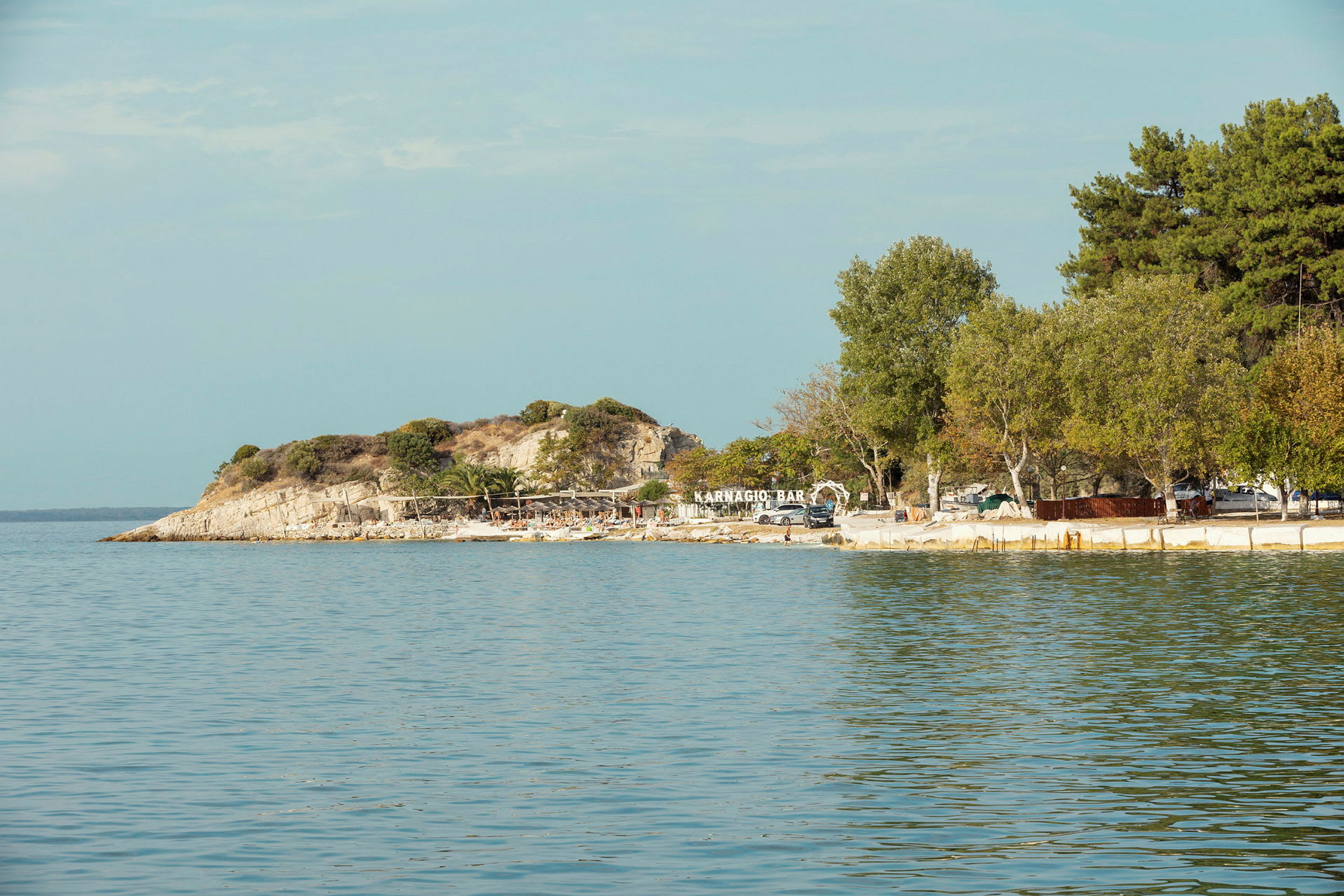 Bild av en kustlinje med en liten strand, träd och en barmarkerad med texten 'Karnagio Bar'. (AI-genererad text)