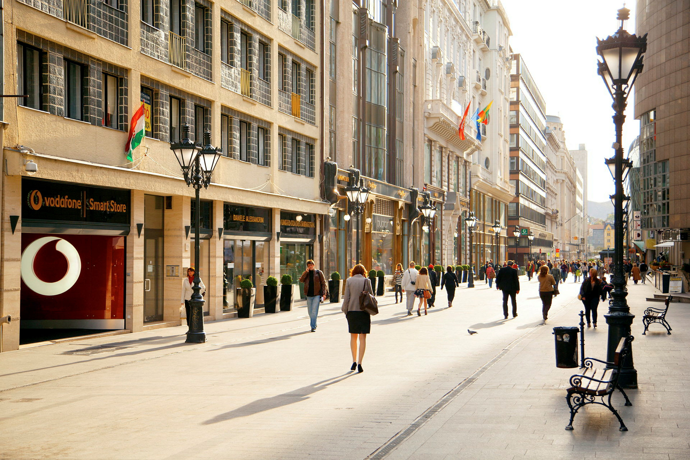 En bred gågata med människor som promenerar, omgiven av butiksfasader och byggnader. (AI-genererad text)