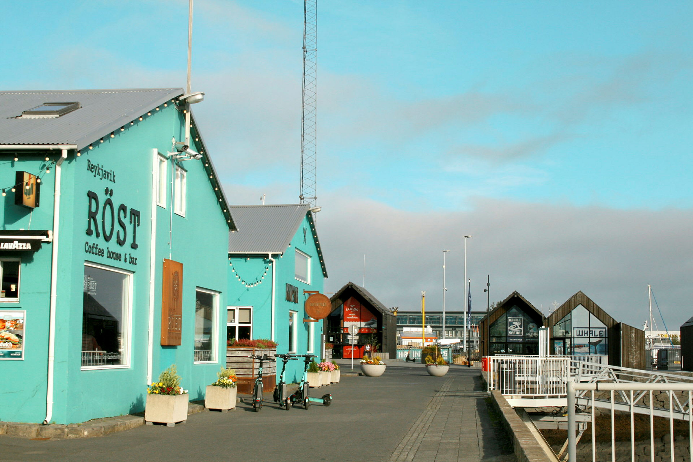 Ett turkost kaféhus vid en hamn med byggnader och trädäck i bakgrunden. (AI-genererad text)