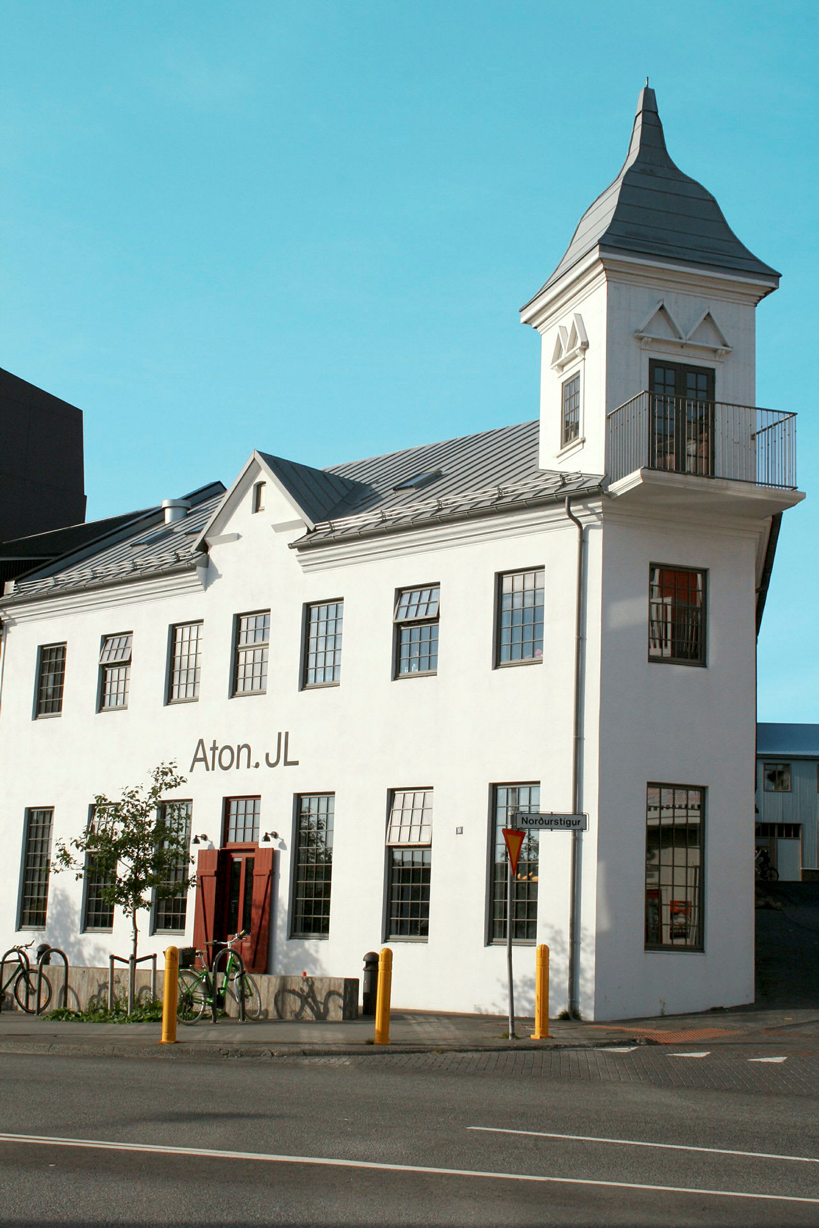 En vit byggnad med ett torn och stora fönster. Byggnaden har en skylt som säger "Aton.JL" och det finns cyklar parkerade framför. (AI-genererad text)
