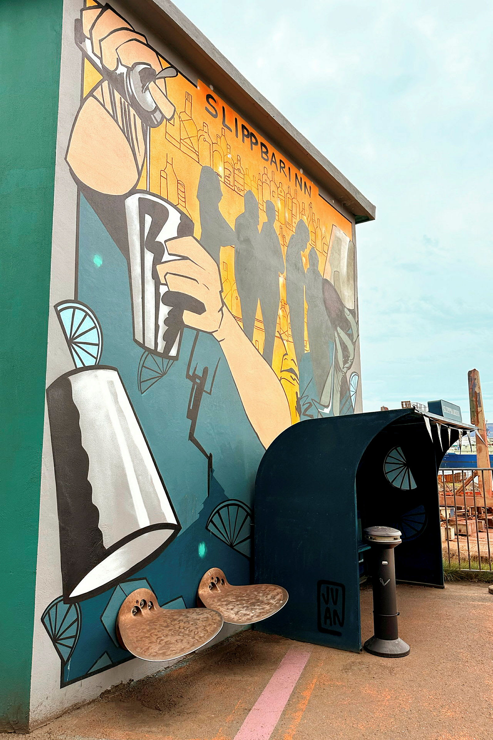 En muralmålning på en vägg bredvid en mörkblå sittplats med en liten avskärmning. Målningen föreställer en bar-miljö med en hand som håller en shaker. (AI-genererad text)