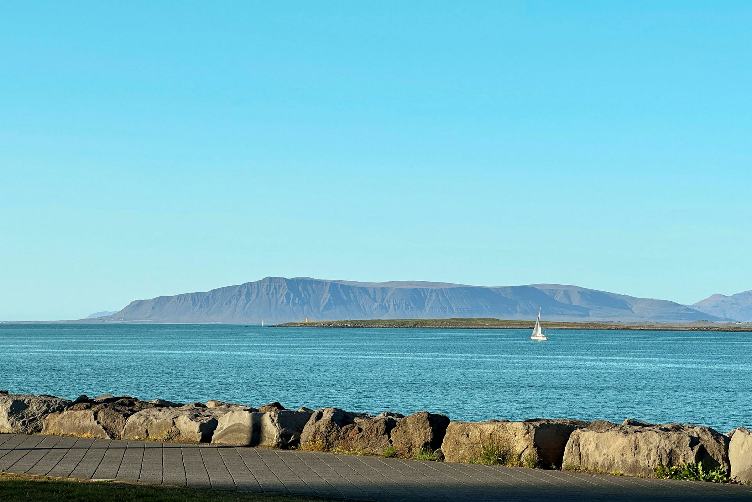 En klippmur vid en kust med hav, en segelbåt och berg i bakgrunden. (AI-genererad text)
