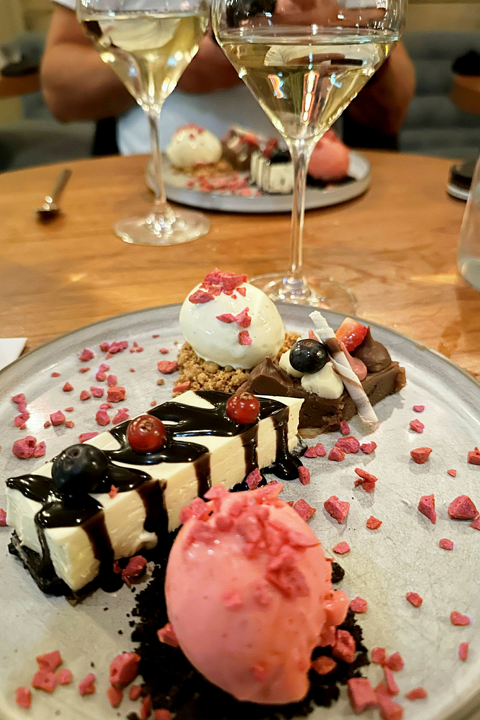 En tallrik med olika desserter, inklusive en rosa sorbet, cheesecake och andra små godsaker, dekorerade med bär och sås. Två glas vitt vin syns i bakgrunden. (AI-genererad text)