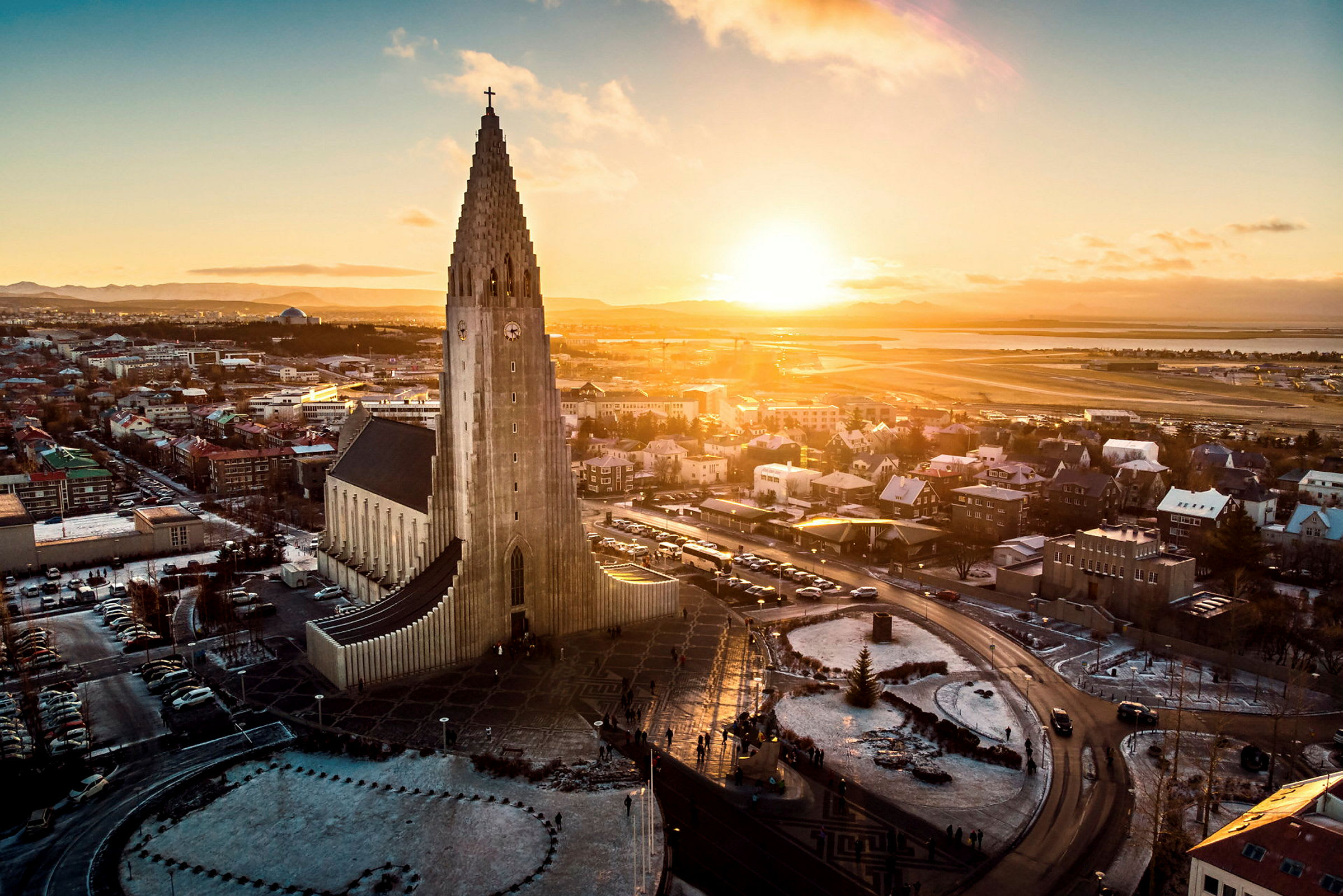 En kyrka med modern arkitektur och en hög tornstruktur, omgiven av en stad och ett landskap under en solnedgång. (AI-genererad text)