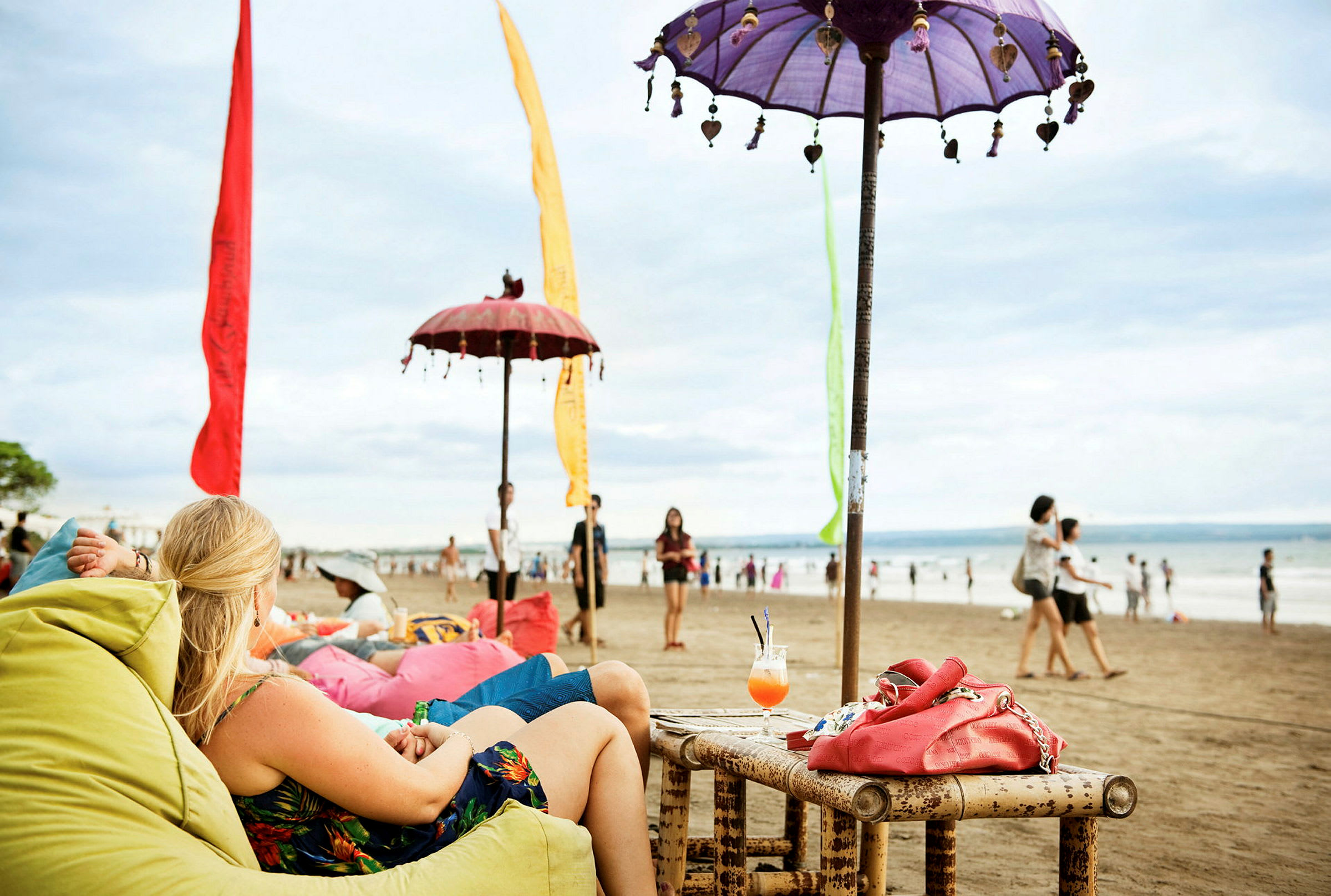 Strand med personer som sitter i färgade säckstolar under dekorerade parasoller. I förgrunden står ett bambubord med en drink och en röd väska. (AI-genererad text)