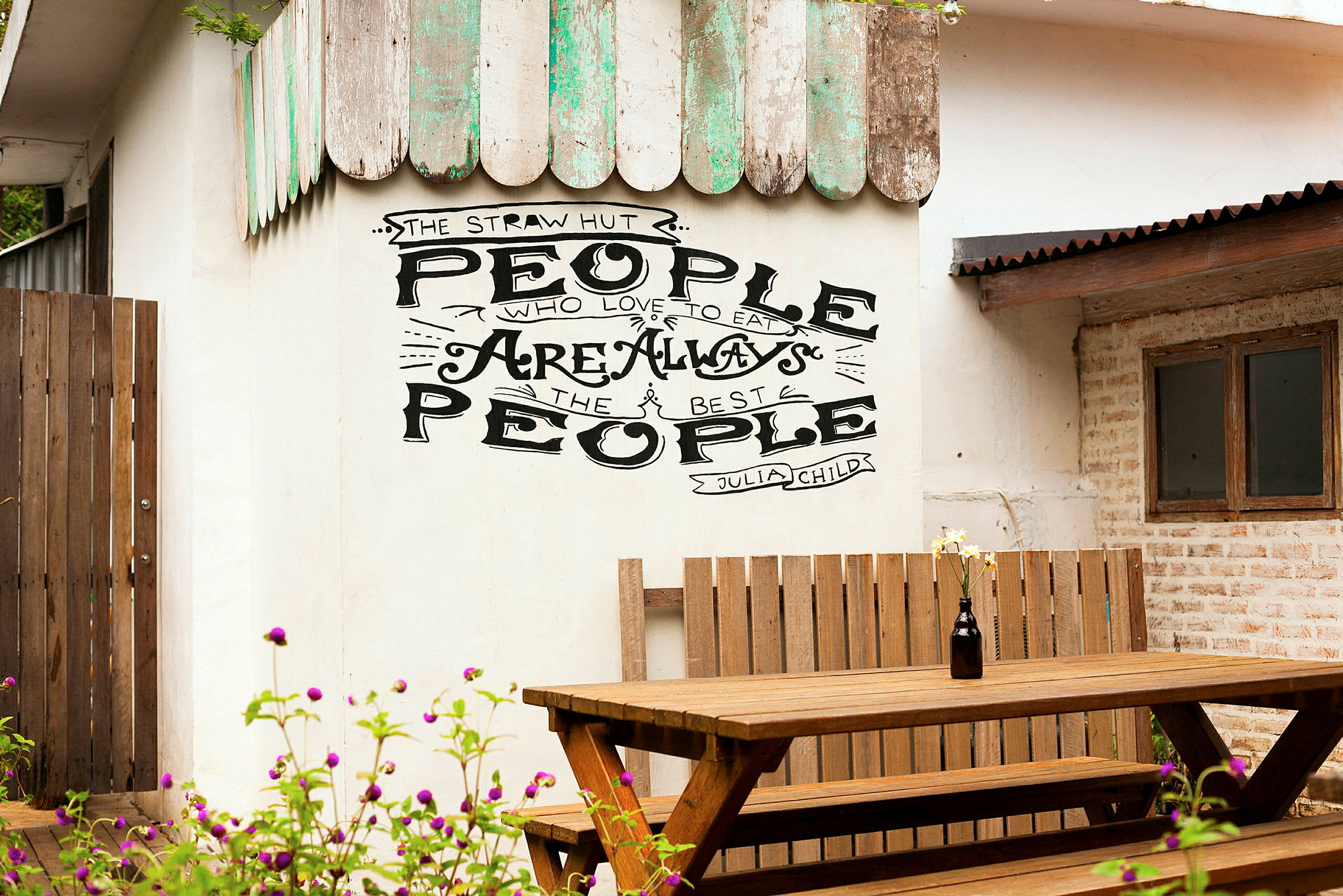 Vägg med texten 'People who love to eat are always the best people' ovanför en träbänk och ett bord. (AI-genererad text)