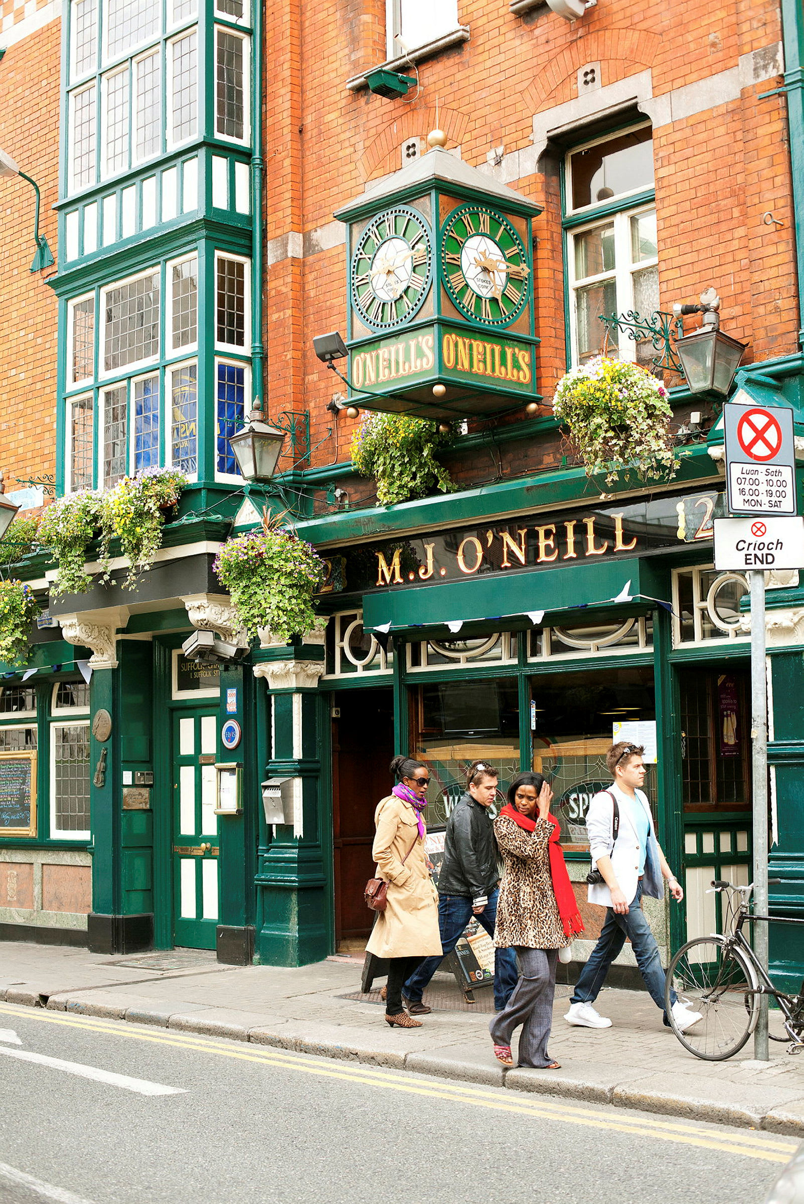 En traditionell irländsk pub med tegelväggar och ett grönt klocktorn ovanför entrén. Flera personer promenerar förbi utanför. (AI-genererad text)