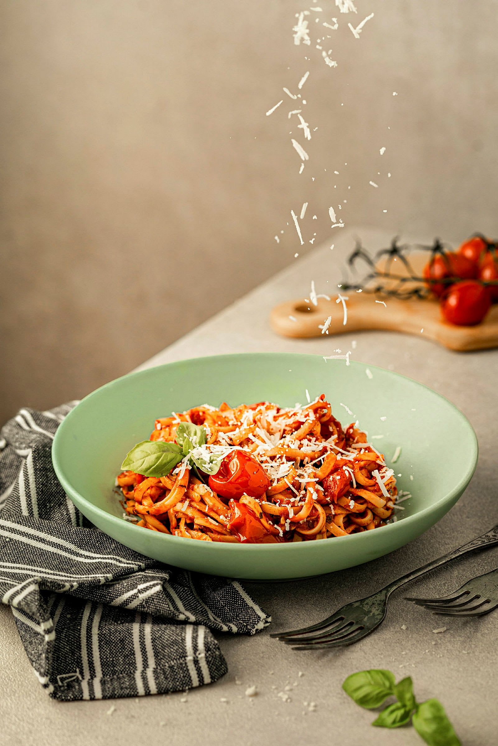 En tallrik med pasta, toppad med tomater, basilika och riven ost, placerad på en grå yta med bestick och en svartvit tygduk. (AI-genererad text)