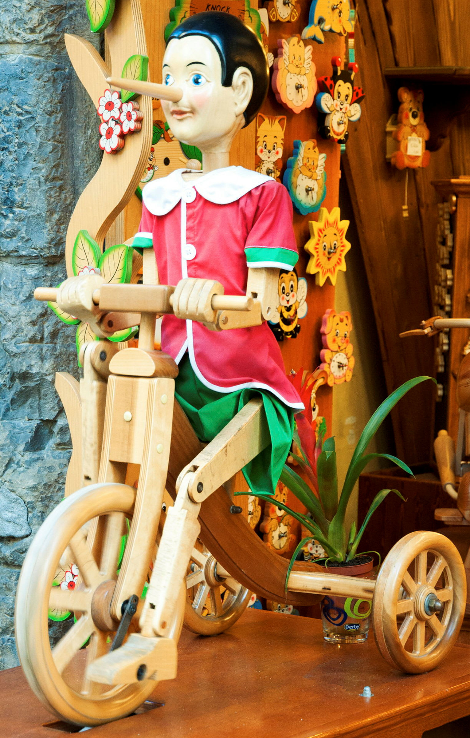 Träfiguren är klädd i färgglada kläder, sittande på en träleksakstricycle framför en dekorativ bakgrund. (AI-genererad text)