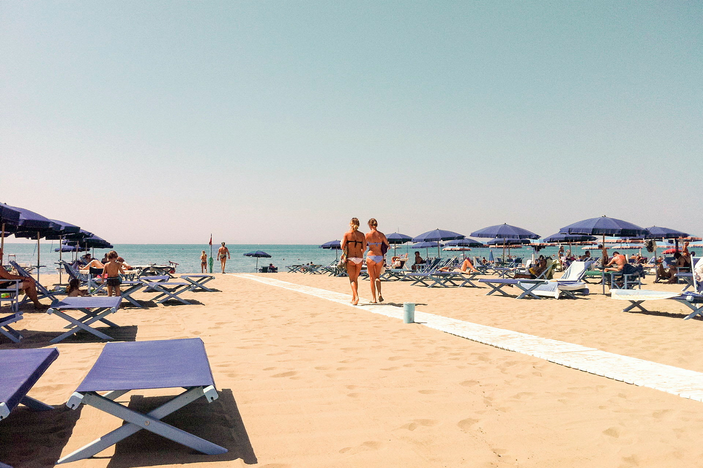 En sandstrand med blå solstolar och parasoll. Människor solar och går på stranden med en klar blå himmel i bakgrunden. (AI-genererad text)