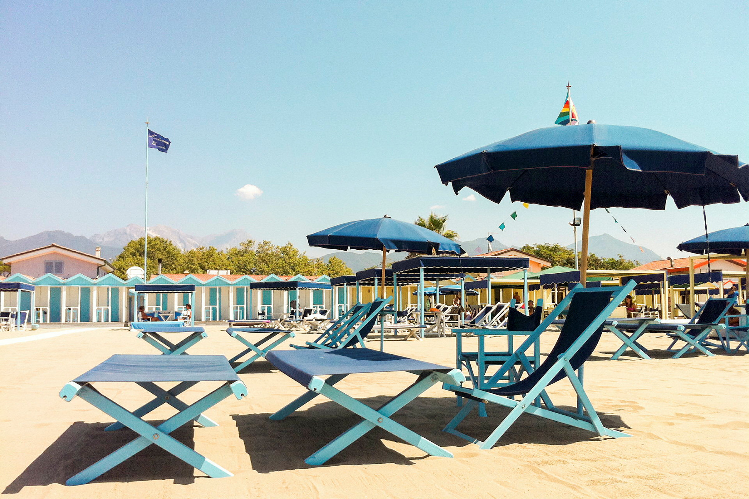 Strandmiljö med solstolar och parasoller samt ett område med strandhyddor i bakgrunden. (AI-genererad text)