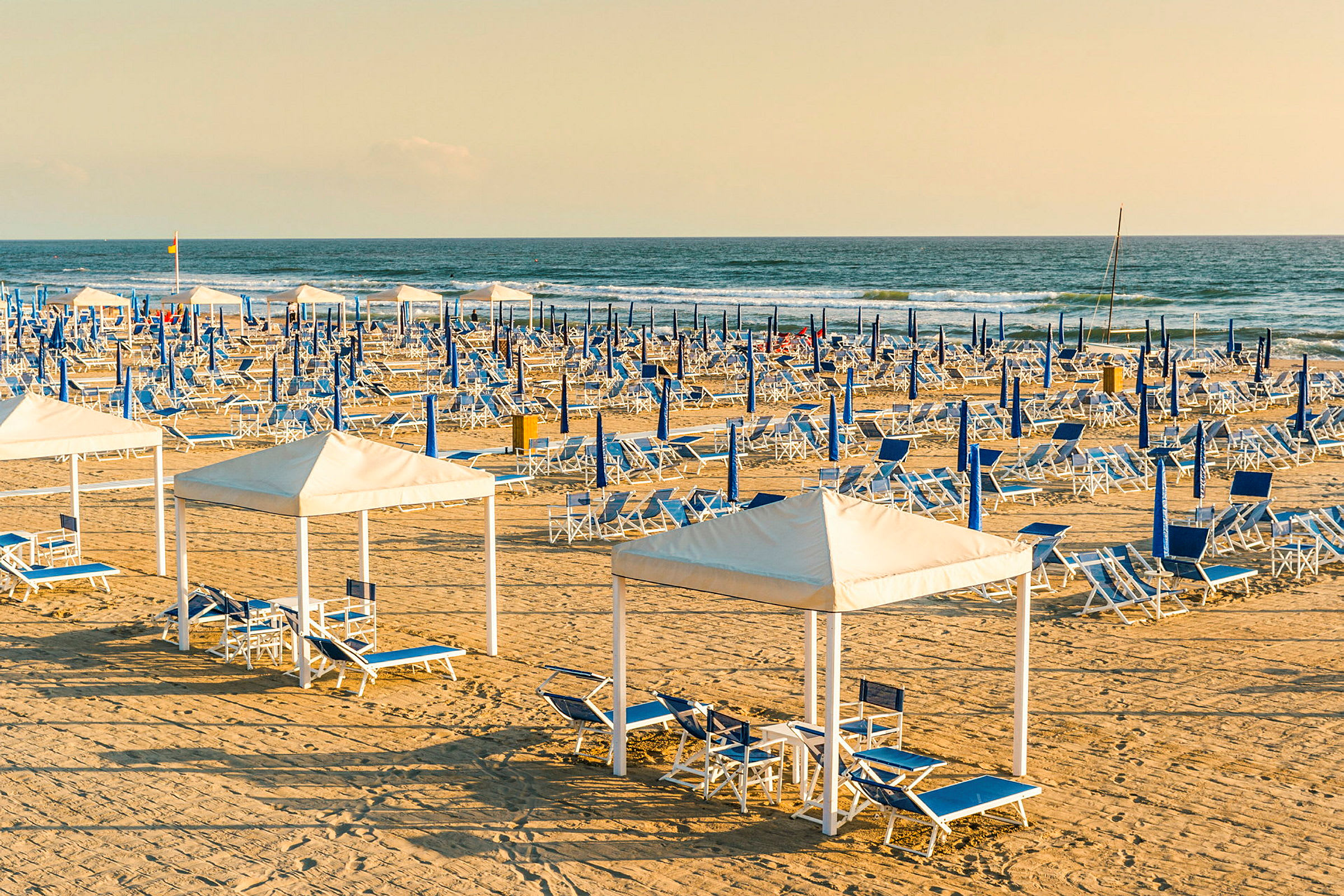 En sandstrand med uppställda blå och vita solstolar samt parasoller. I förgrunden finns cabanas med sittgrupper. (AI-genererad text)