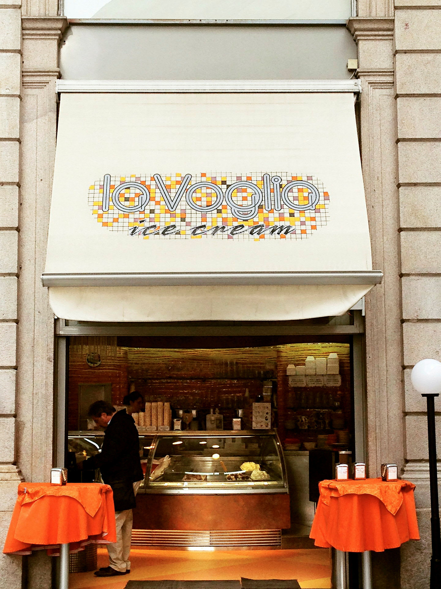 En glassbutik med skylt som säger "La Voglia ice cream" och två små bord med orange dukar framför ingången. (AI-genererad text)