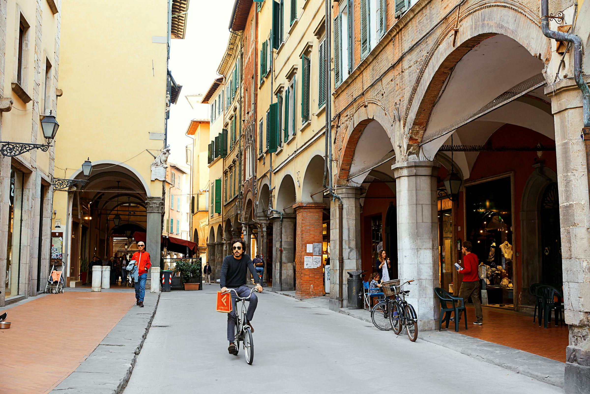 Gata med historiska byggnader och arkader. En person cyklar förbi, och några personer promenerar eller sitter vid ett kafé. (AI-genererad text)