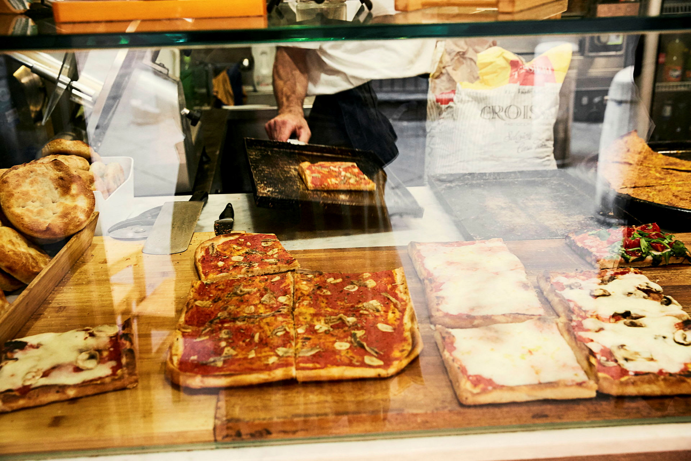 Rutor av pizza med olika tillbehör bakom en glasdisk, samt bröd och en pizzaugn i bakgrunden. (AI-genererad text)