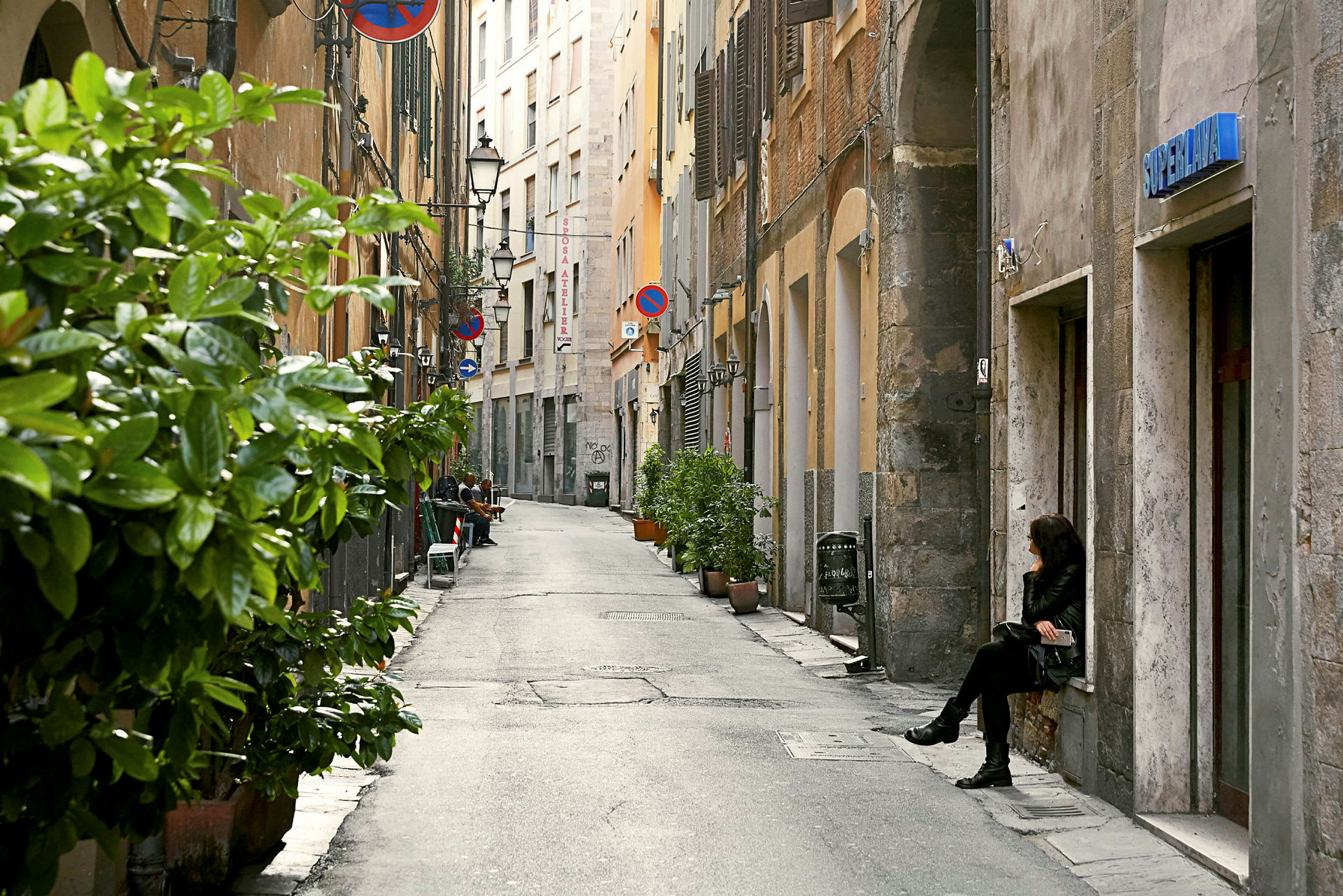 En smal gata med byggnader på båda sidor, några krukväxter längs trottoaren och en person som sitter vid väggen till höger. (AI-genererad text)