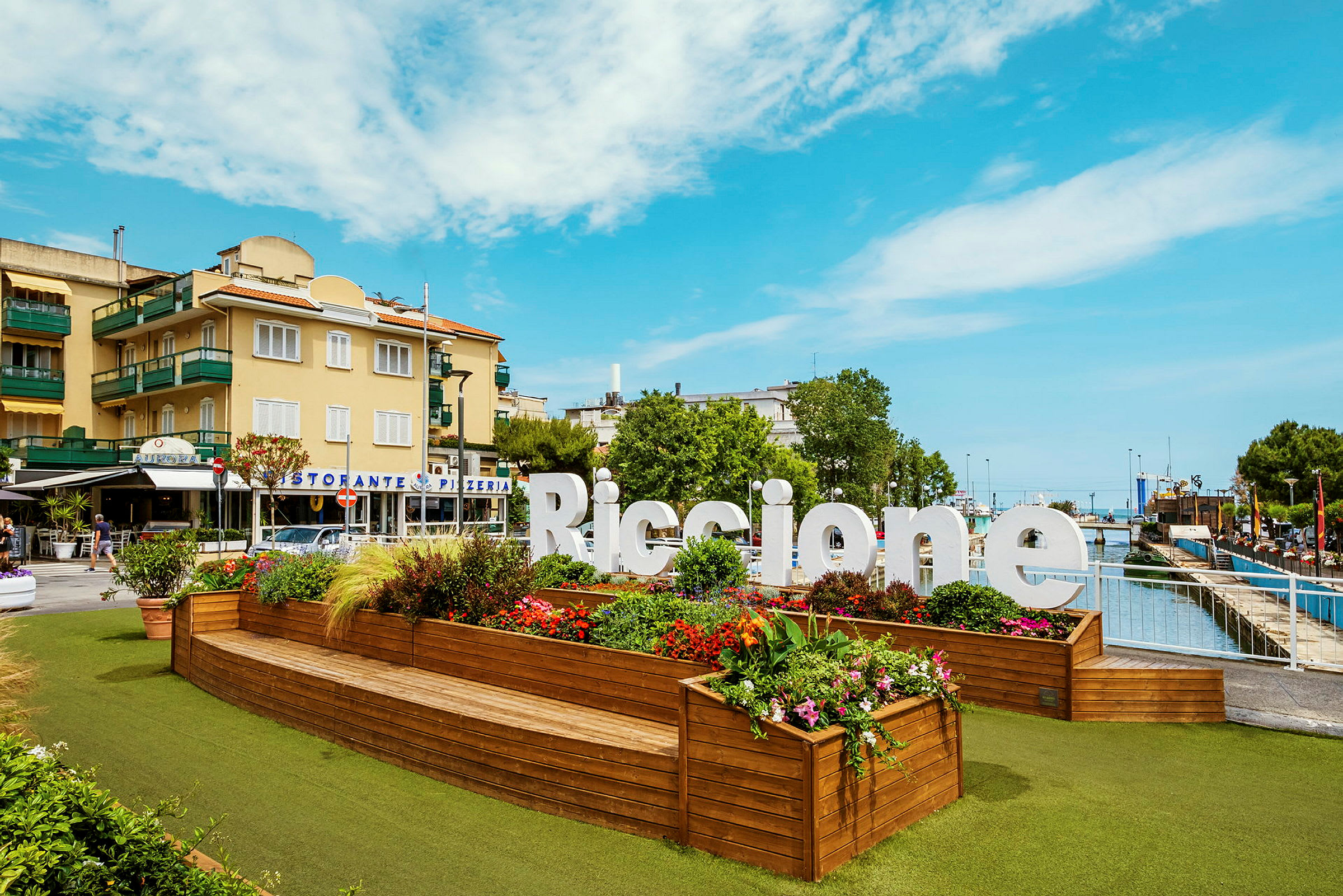En stor skylt med texten "Riccione" står framför ett planteringsområde med färgglada blommor och gröna växter. I bakgrunden syns byggnader och en klar himmel. (AI-genererad text)