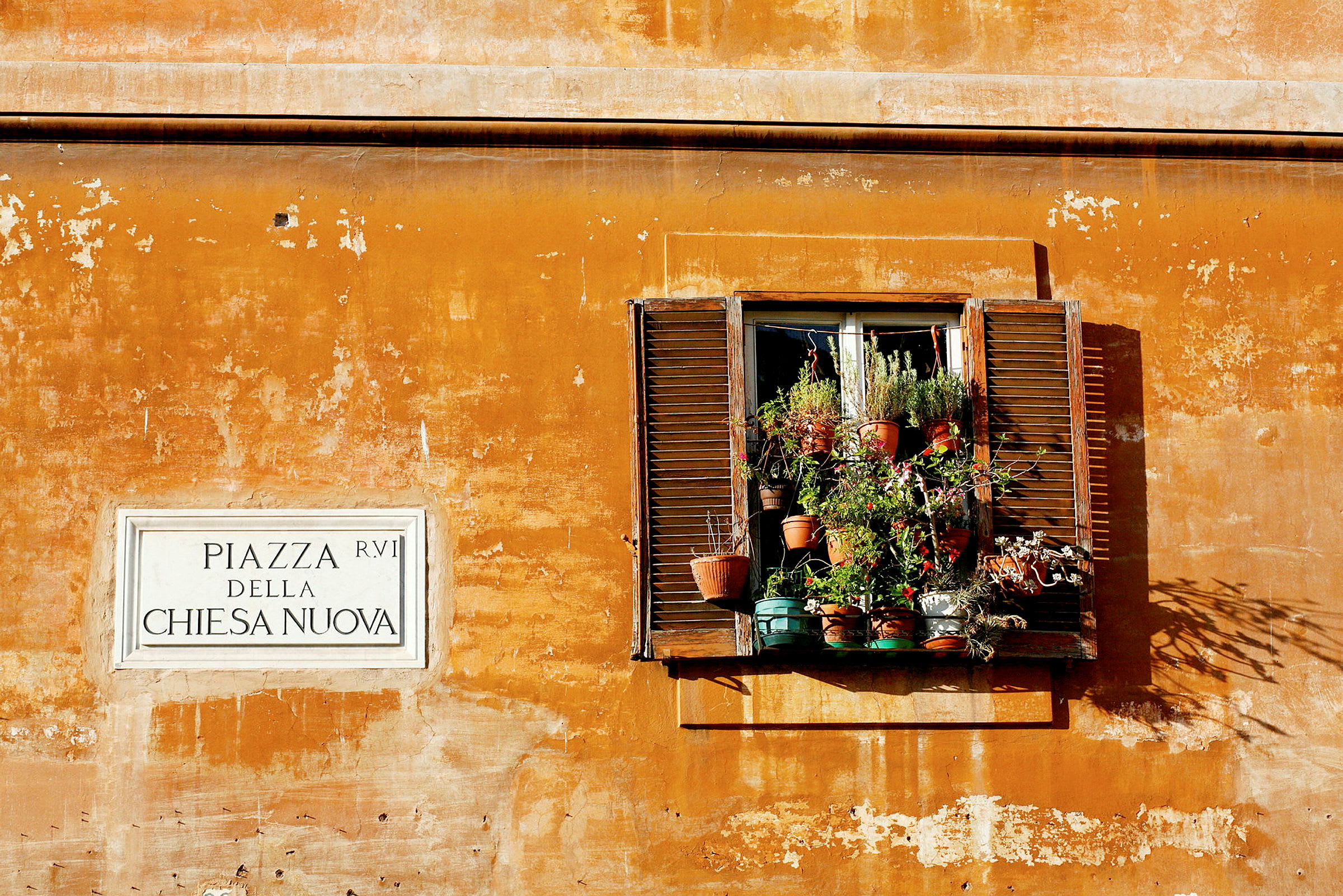 Ett fönster med planteringskrukor på en gulmålad vägg, bredvid en skylt med texten 'Piazza della Chiesa Nuova'. (AI-genererad text)