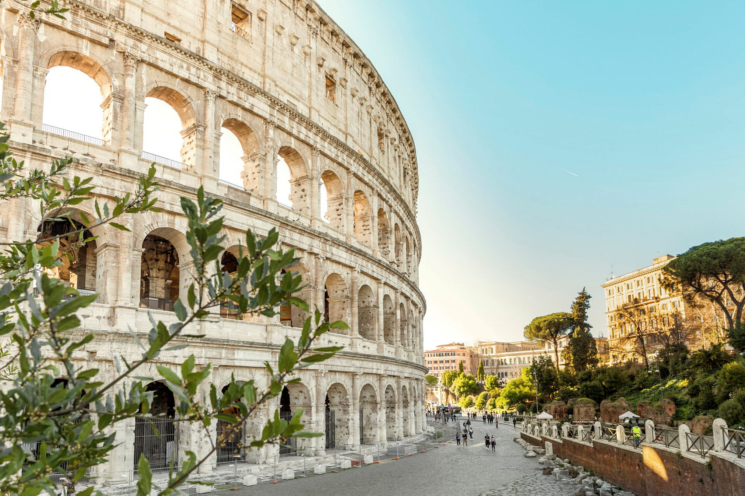 Bild av Colosseum med omgivande romerska arkitektur och grönska under en klar blå himmel. (AI-genererad text)