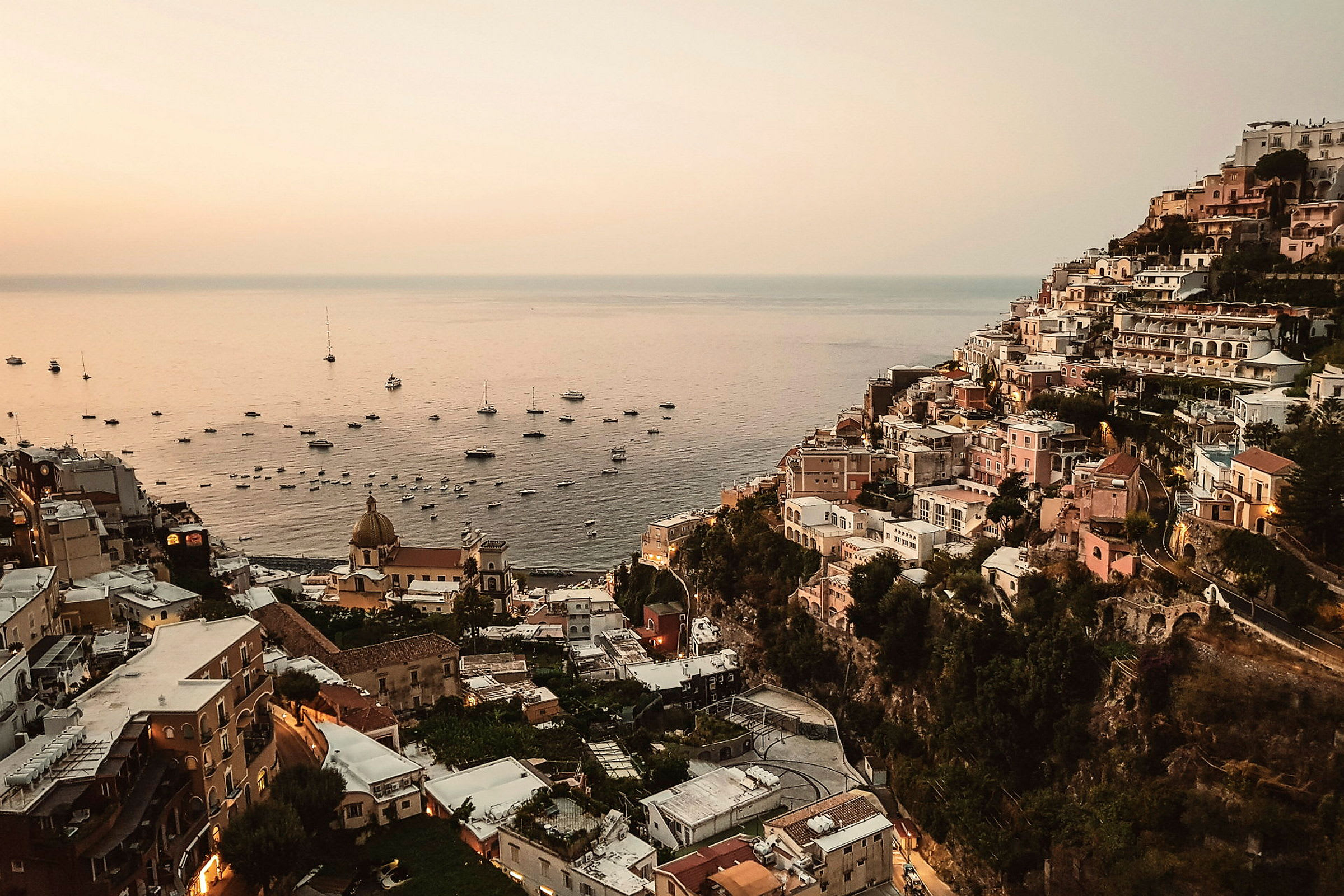 Bild av Positano under solnedgång med byggnader längs en sluttning och havet fullt av båtar. (AI-genererad text)