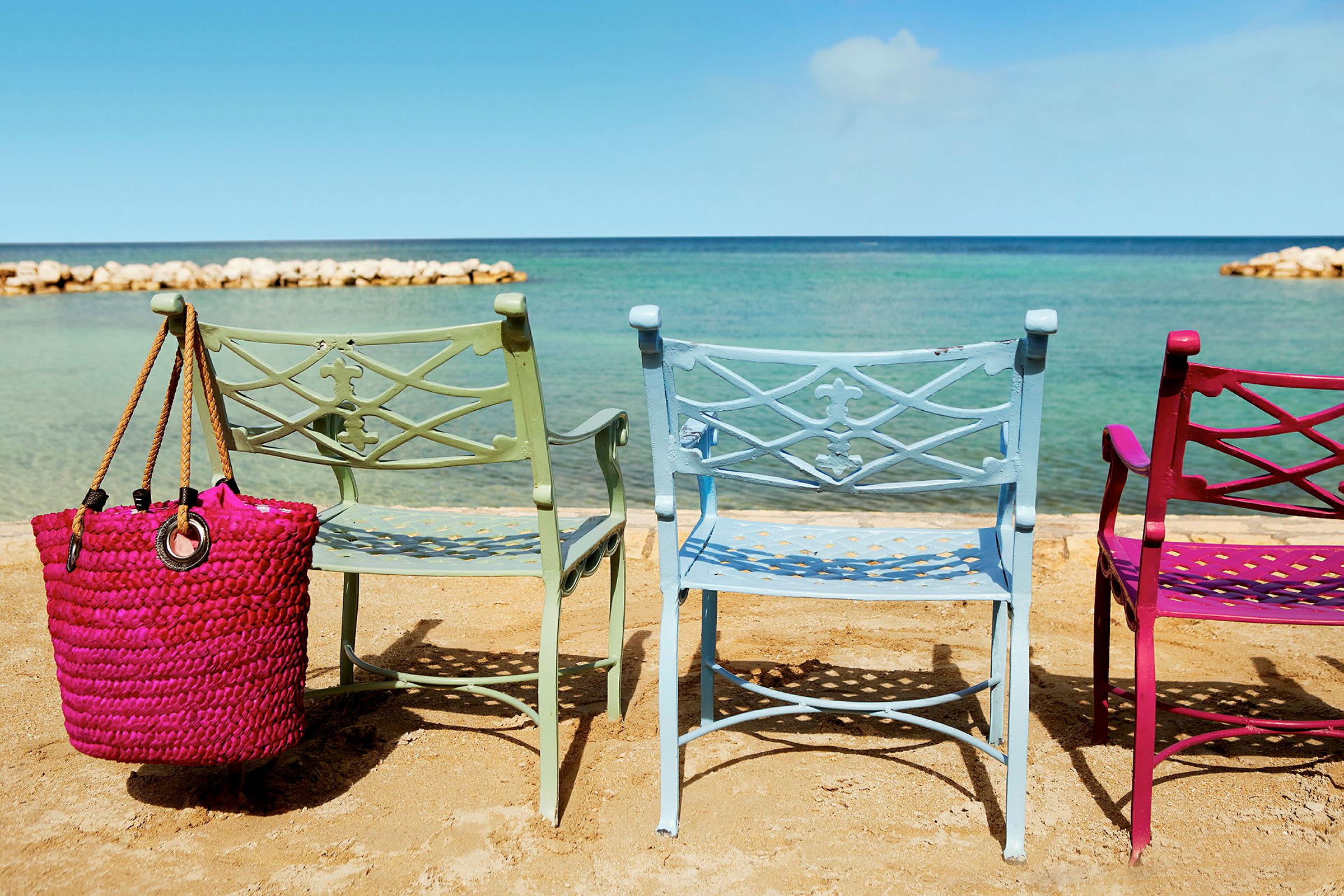 Tre färgglada stolar står på en strand med havet i bakgrunden. En rosa strandväska hänger från en av stolarna. (AI-genererad text)