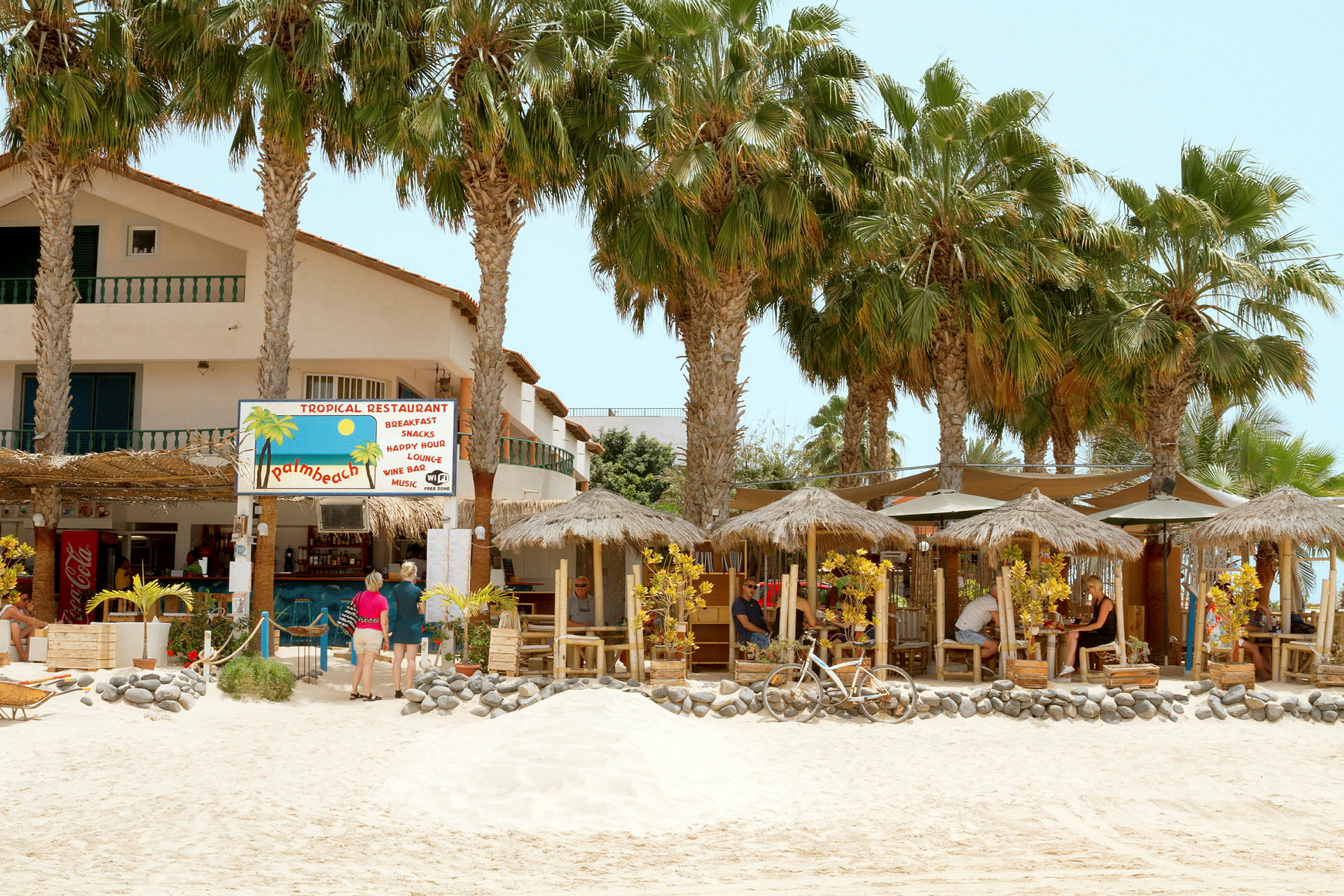 En tropisk restaurang med halmtak och palmer på en sandstrand. (AI-genererad text)