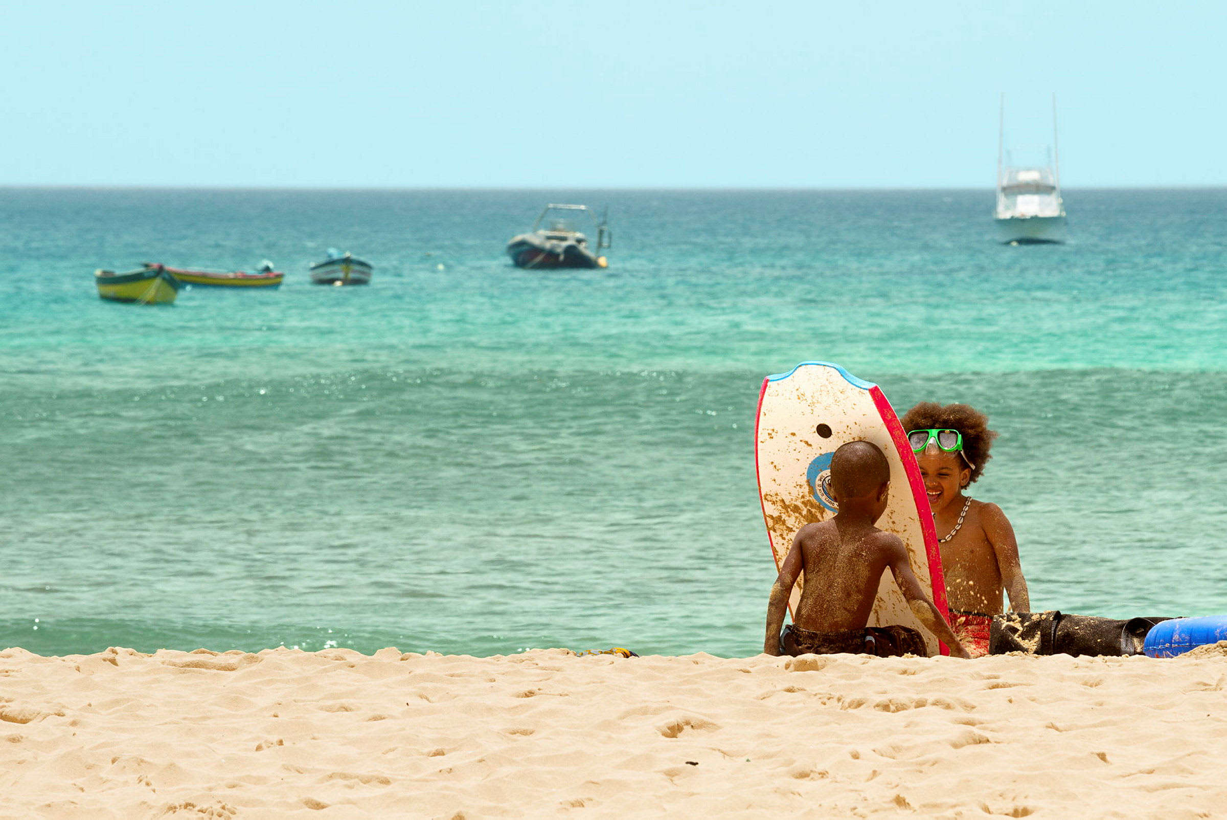 Två personer sitter på en sandstrand med en skimboard i närheten, havet och båtar syns i bakgrunden. (AI-genererad text)