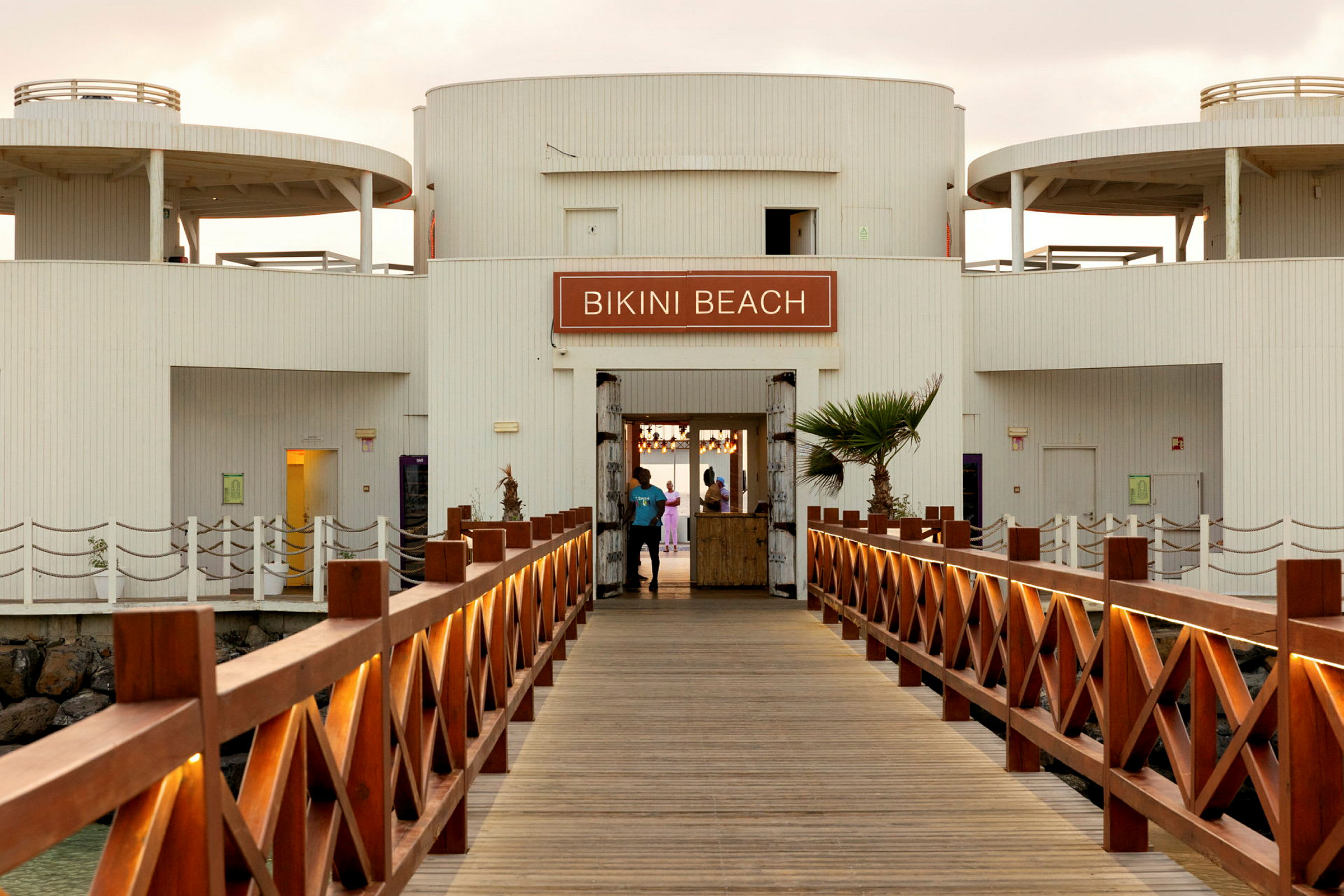 Vy av en träbrygga som leder till en byggnad med skylten 'Bikini Beach'. (AI-genererad text)