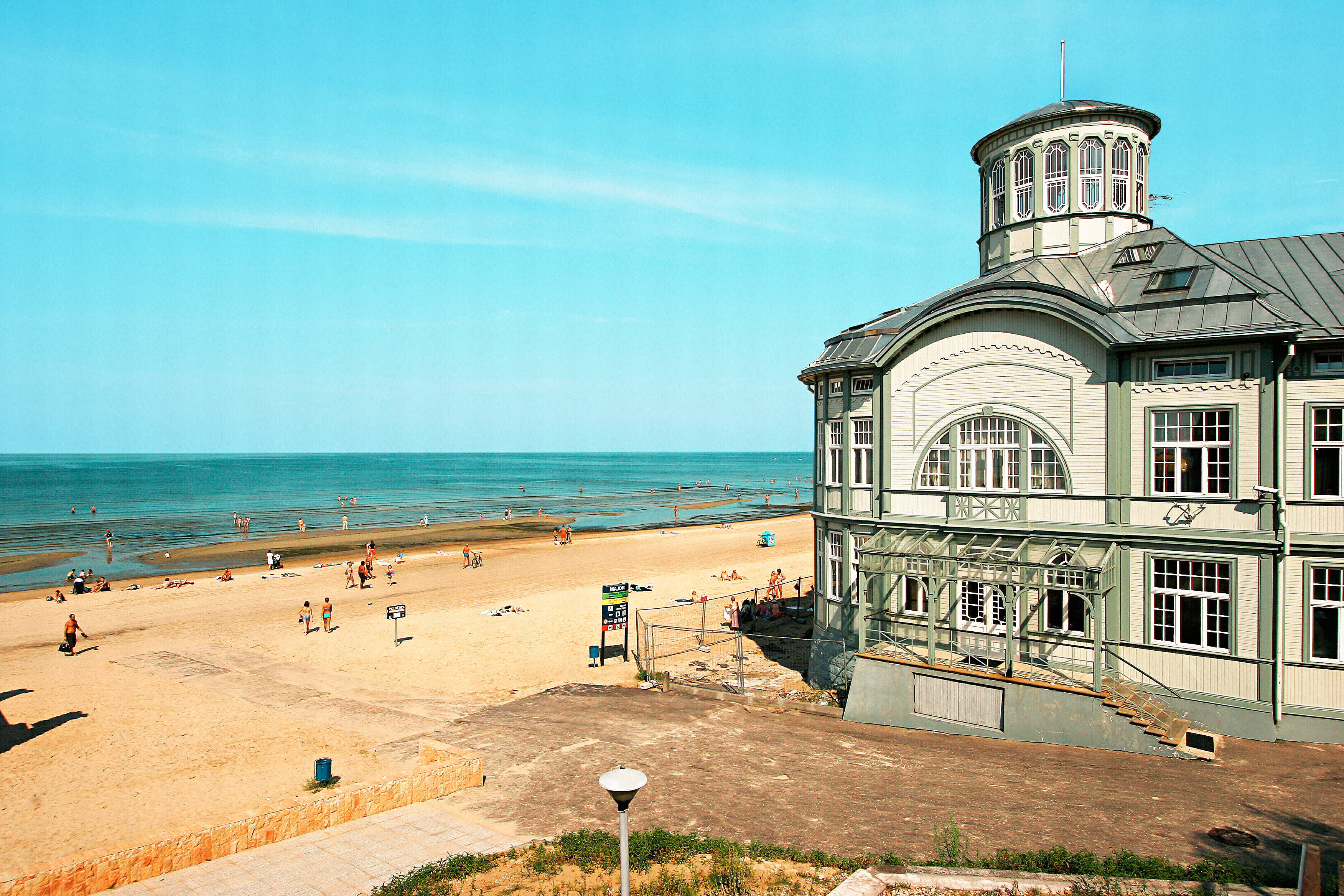 En strand med solbadande människor vid en lugn kustlinje, samt en större byggnad med klassisk arkitektur intill stranden. (AI-genererad text)