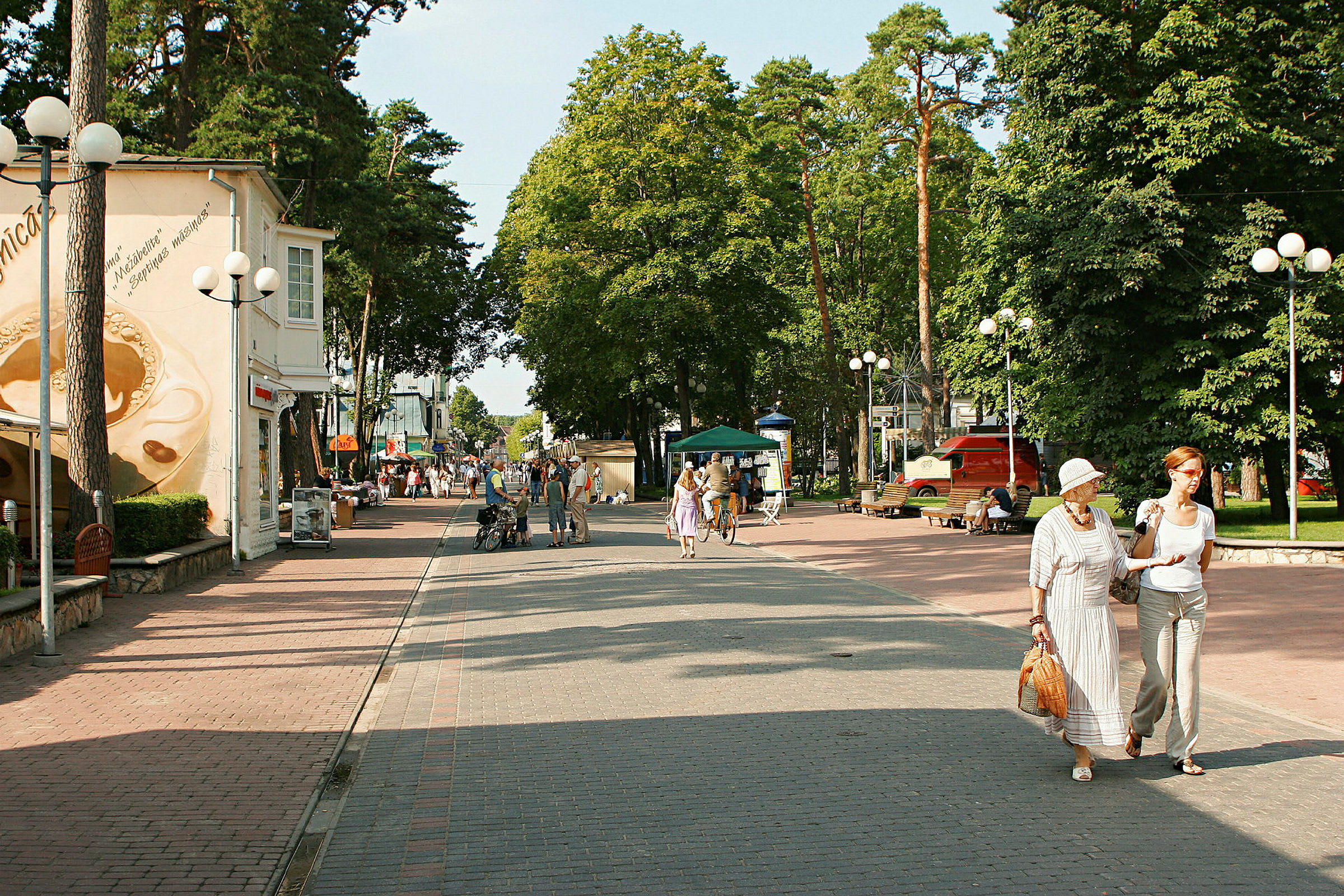 En gågata med människor, omgivande hus, träd och parkeringsområden. (AI-genererad text)