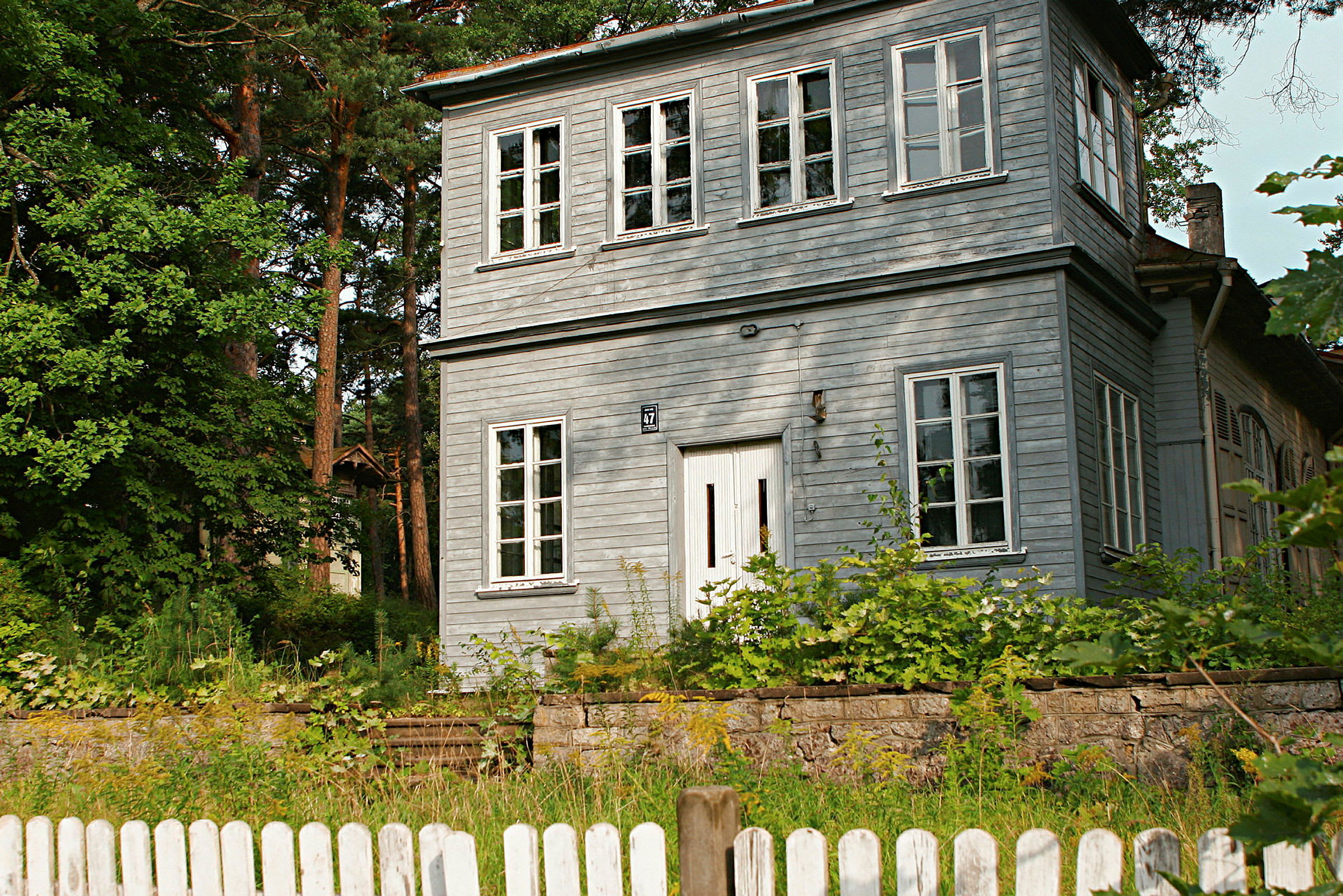 En tvåvånings trähus målat i grå färg, omgiven av grönska och med ett vitt staket i förgrunden. (AI-genererad text)