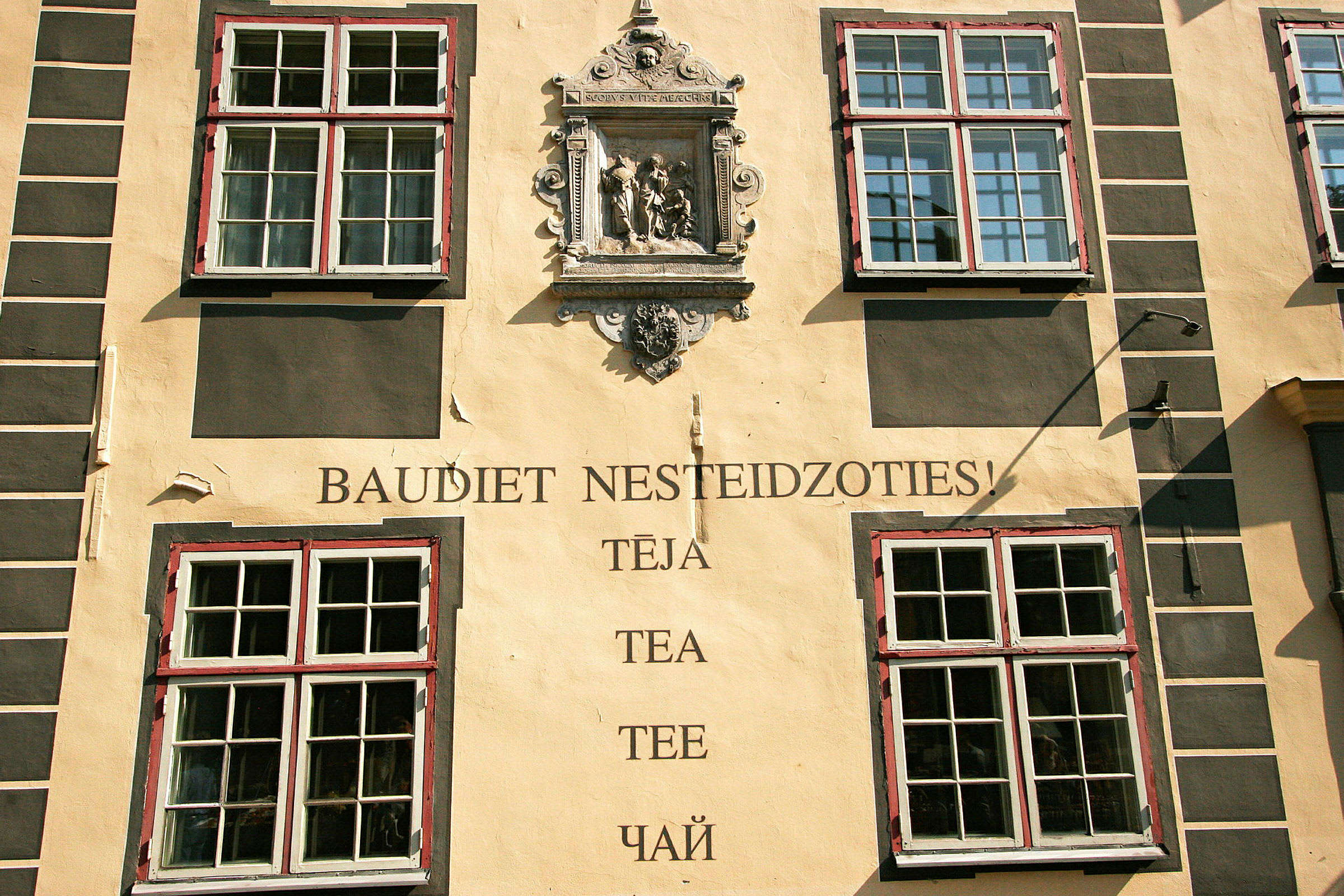 Fasad av en byggnad med fyra stora fönster, en dekorativ stenrelief och texten "Baudiet nesteidzoties! Tēja, Tea, Tee, Чай". (AI-genererad text)