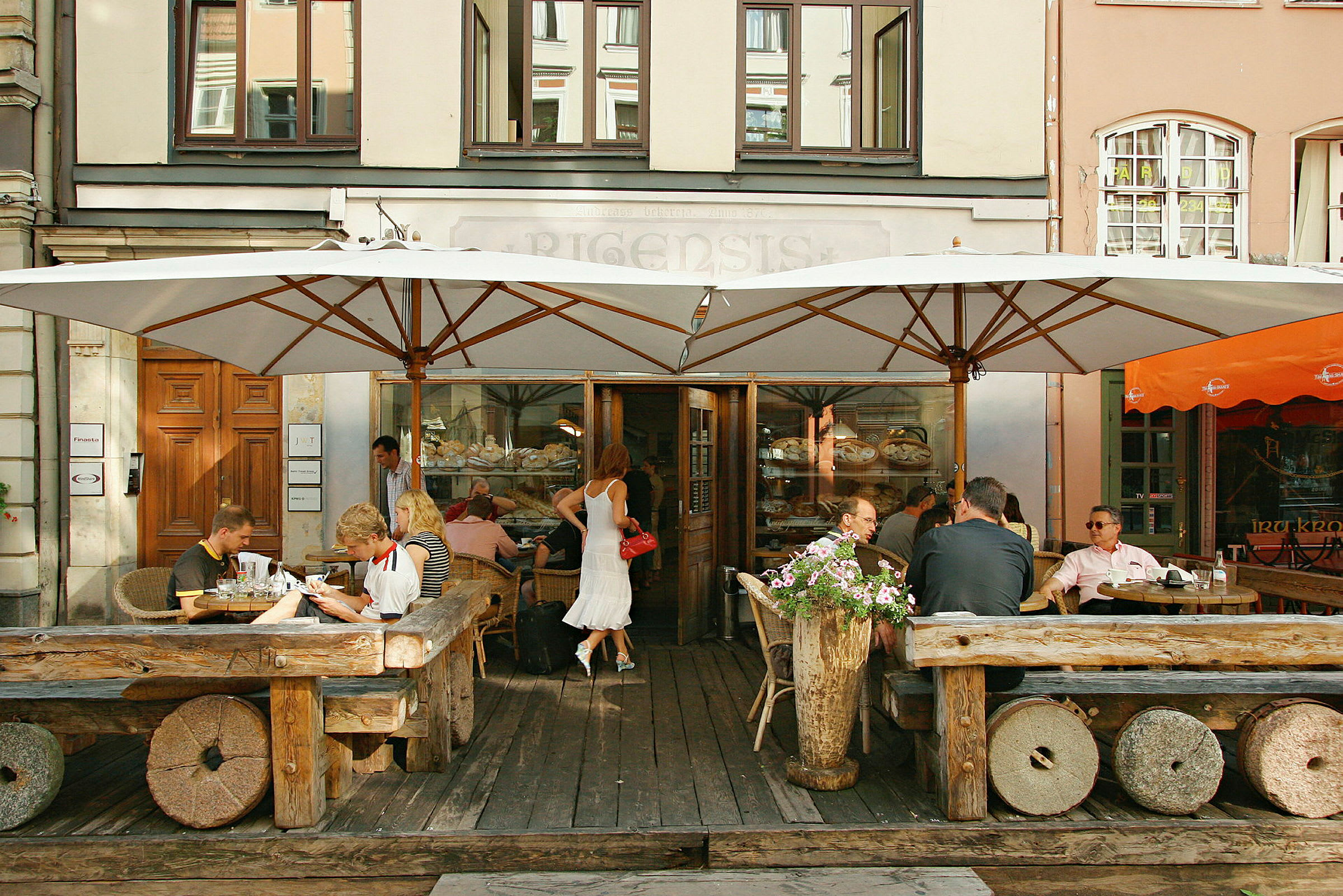 Ett kafé med gäster som sitter ute vid rustika träbord under stora parasoller. (AI-genererad text)