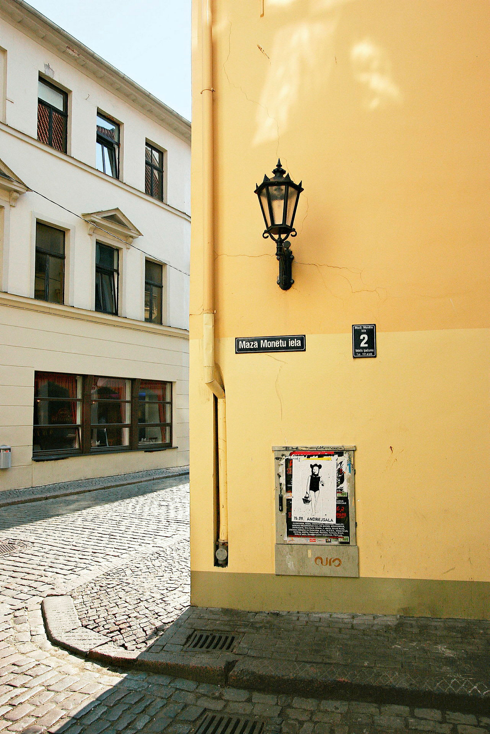 En hörna av en byggnad med gul fasad, en gatlykta, en skylt som anger gatan 'Maza Monētu iela', och ett affischområde. (AI-genererad text)