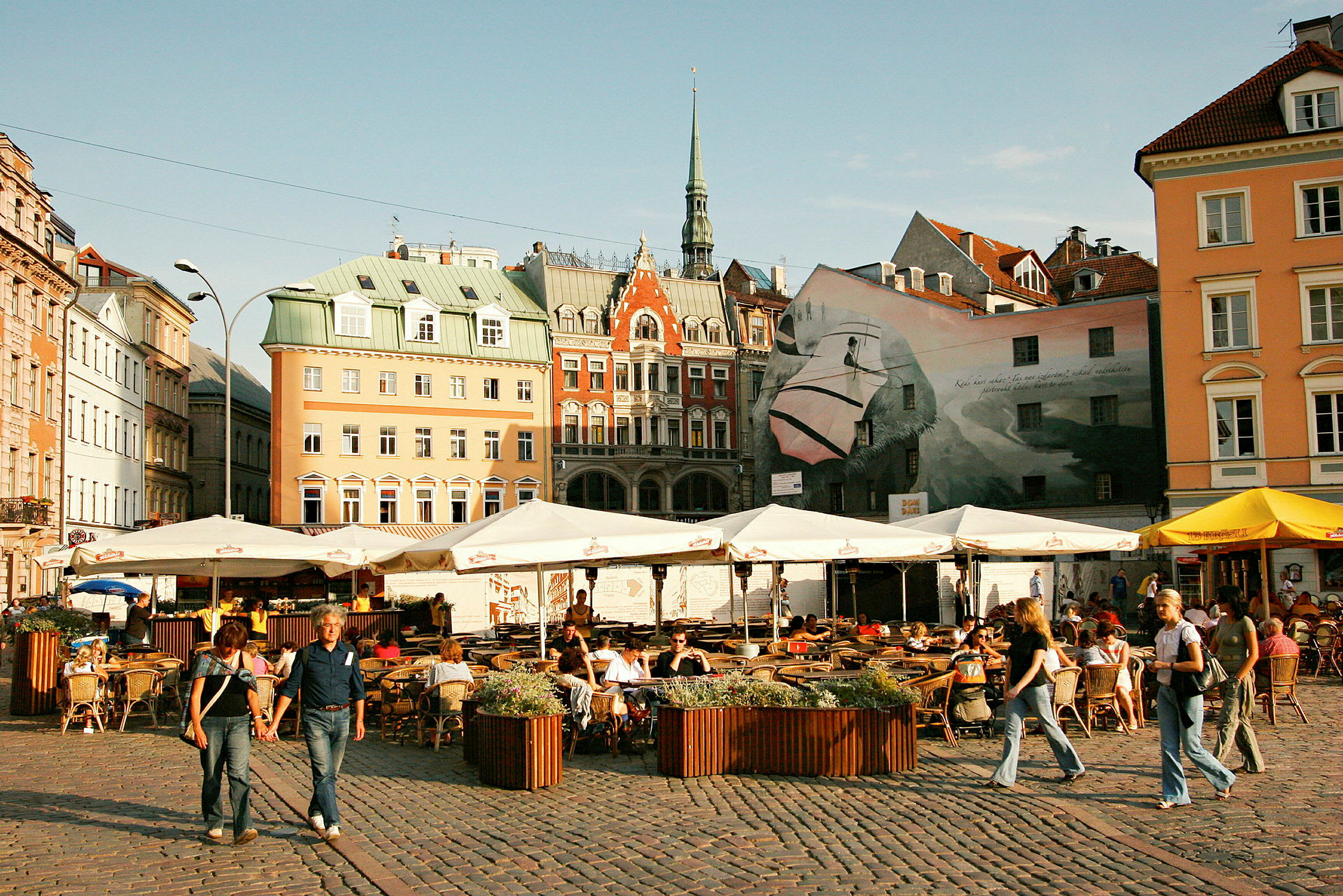 Ett torg med uteserveringar omgivna av äldre byggnader och en stor väggmålning. (AI-genererad text)
