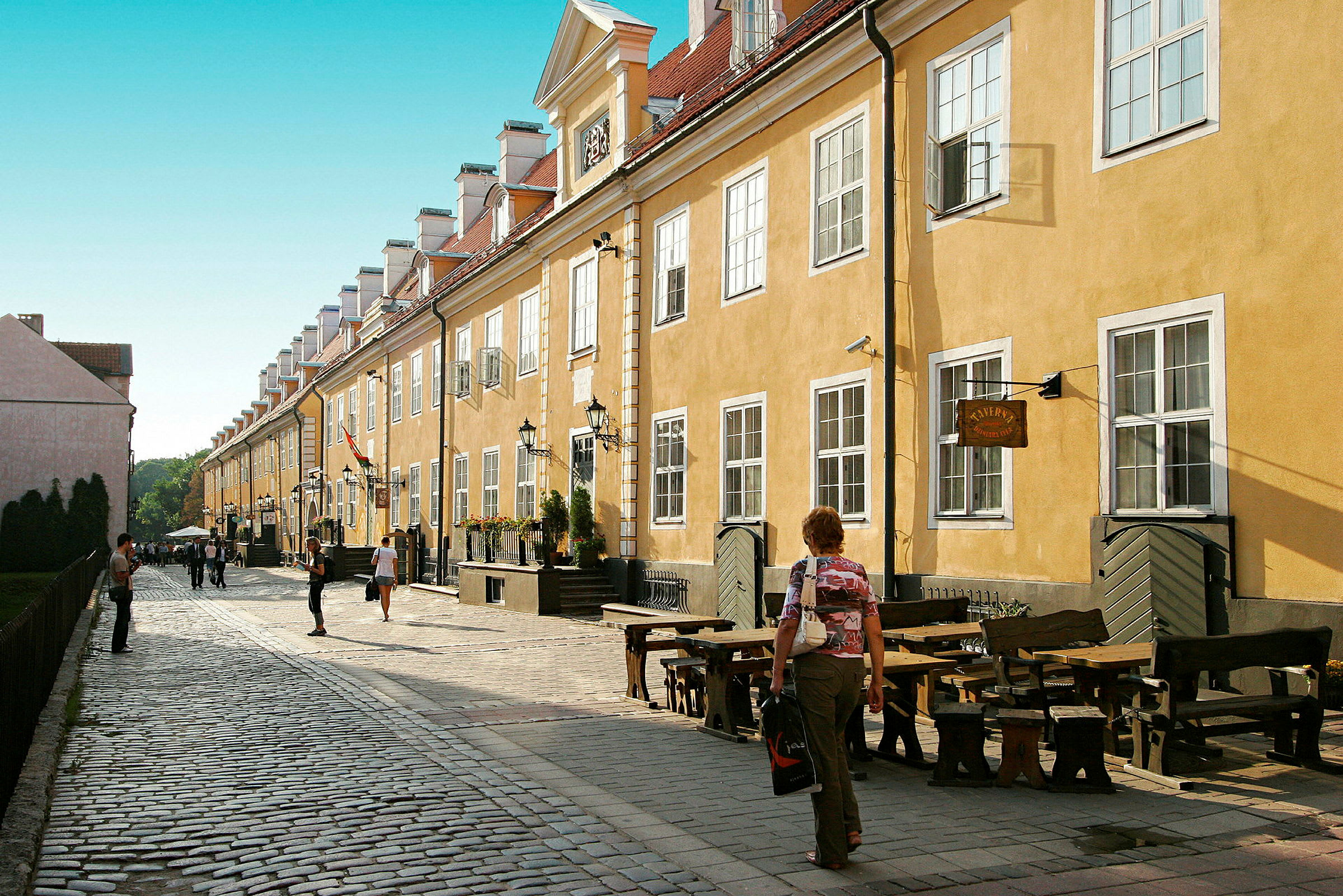 En kullerstensgata kantad av gula byggnader med vita fönster och några uteserveringar. (AI-genererad text)