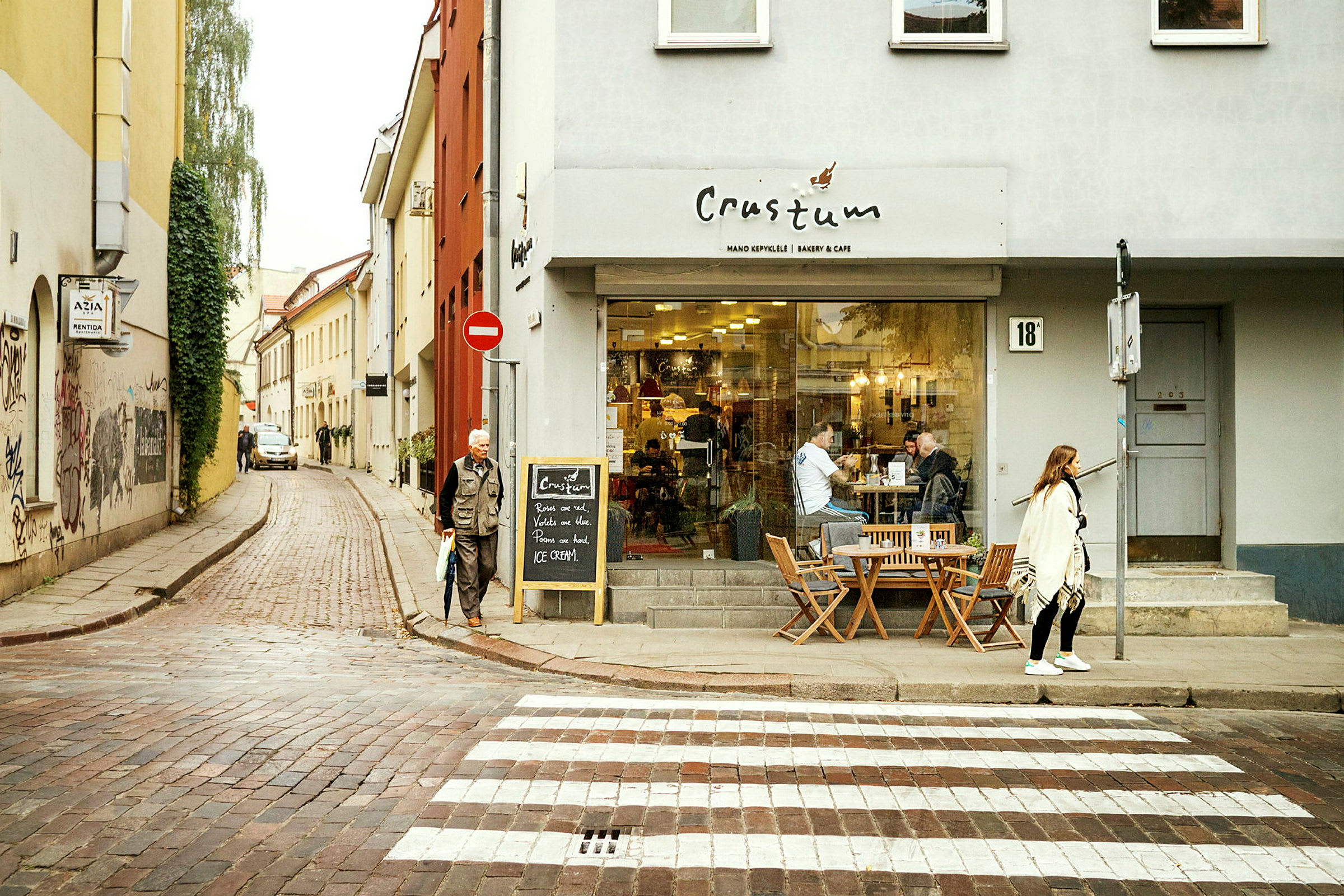 En gata med ett café på högra sidan. Människor sitter utomhus på caféets uteservering och en person går över ett övergångsställe. (AI-genererad text)