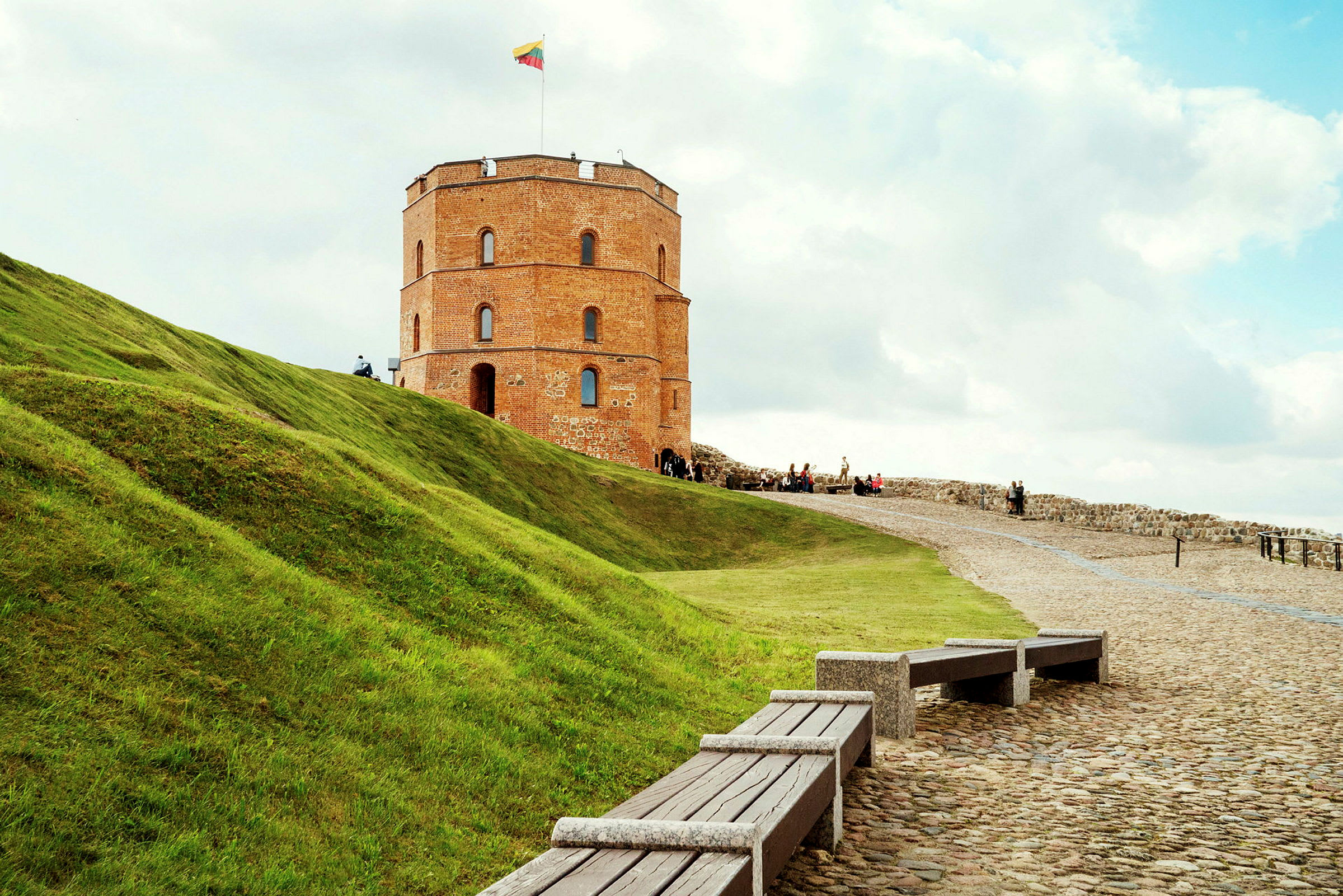 En gammal tegelbyggnad med ett runt torn står på en kulle med grön gräsmatta. På tornet vajar en litauisk flagga. (AI-genererad text)