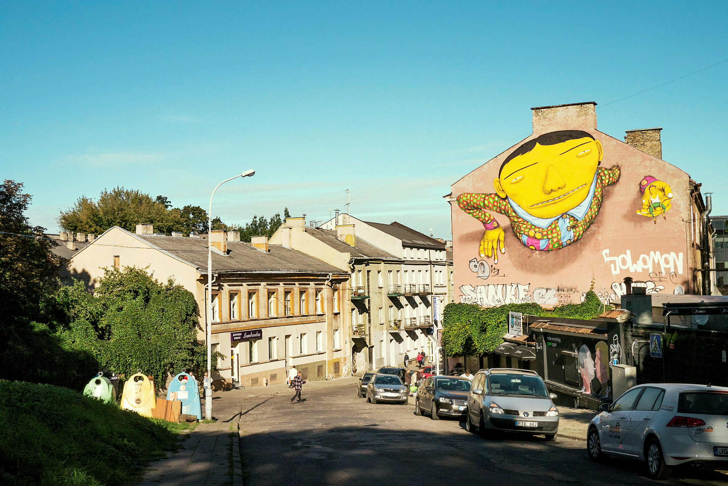 En stadsgata med byggnader och en stor väggmålning föreställande en gul figur. Bilar är parkerade längs gatan. (AI-genererad text)