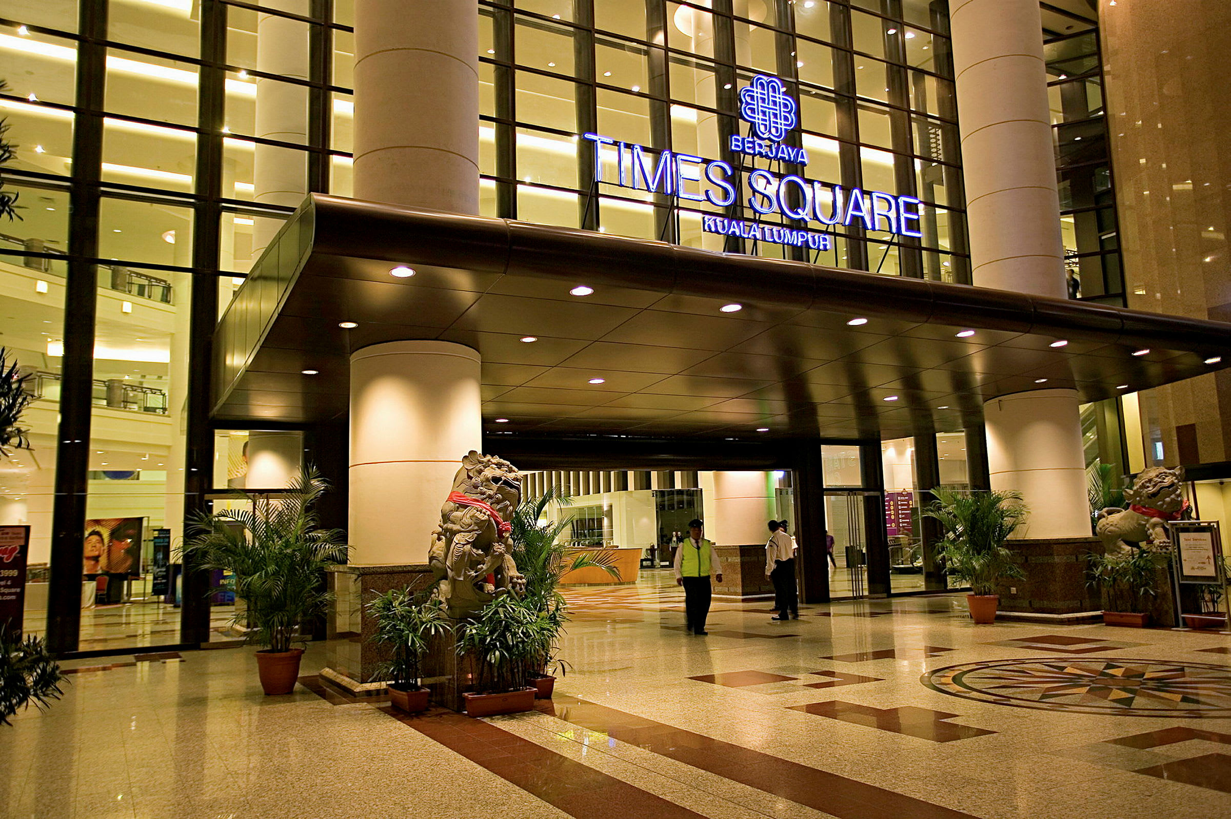 Entréområde till en byggnad med glasfasad och ljus belysning. En skylt över entrén anger 'Berjaya Times Square Kuala Lumpur'. Trägolv och dekorativa växter syns också. (AI-genererad text)
