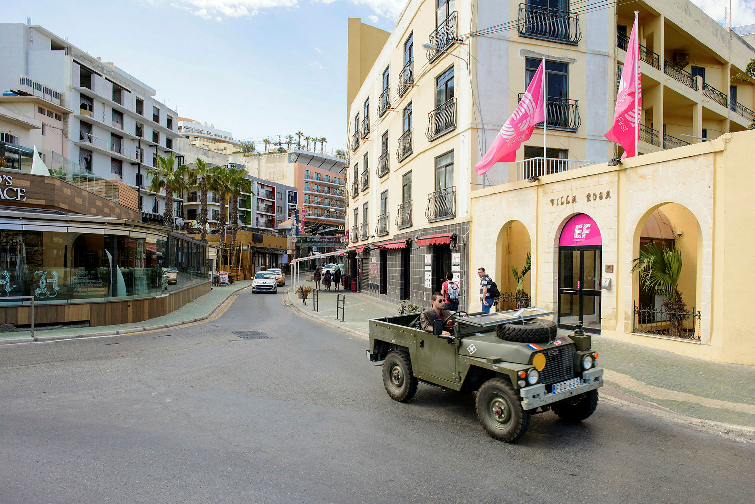 En gata med byggnader, inklusive en gul fasad med rosa flaggor och en butik med glasfönster. En militärgrön jeep är parkerad vid vägkanten. (AI-genererad text)