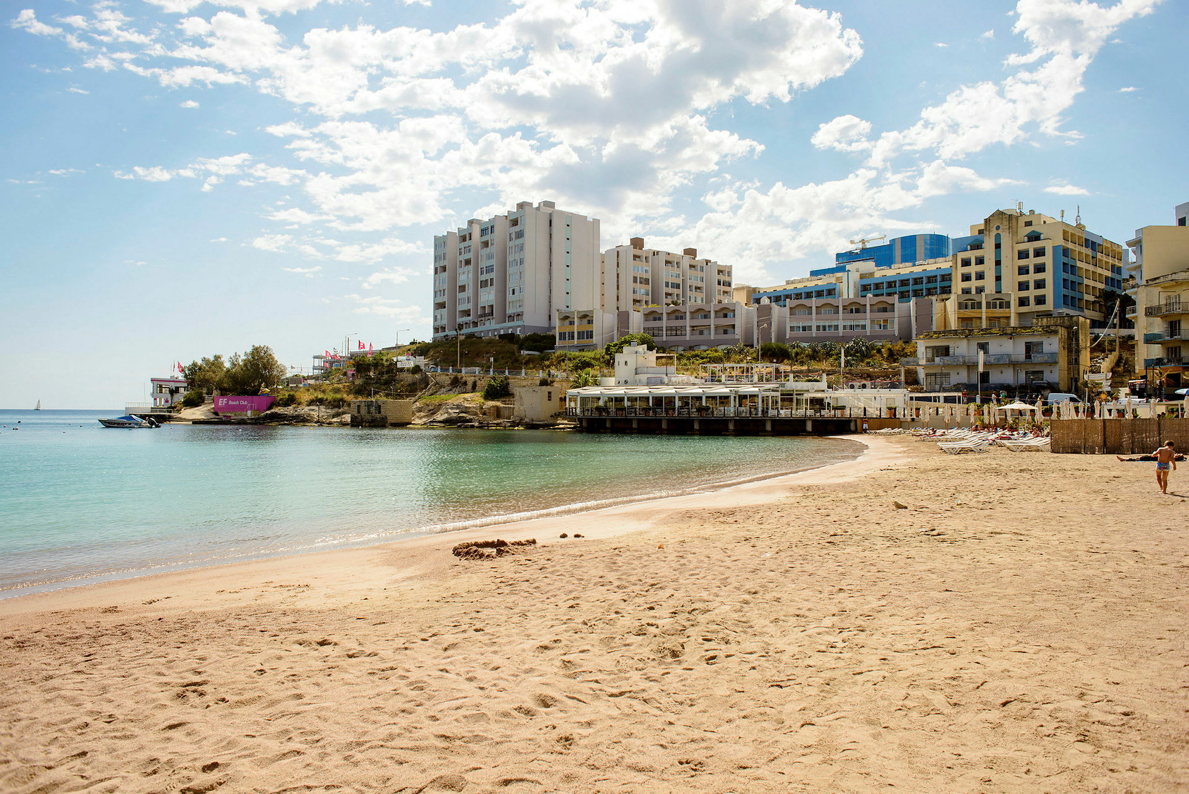 Sandstrand vid stilla havsvatten med byggnader i bakgrunden. (AI-genererad text)