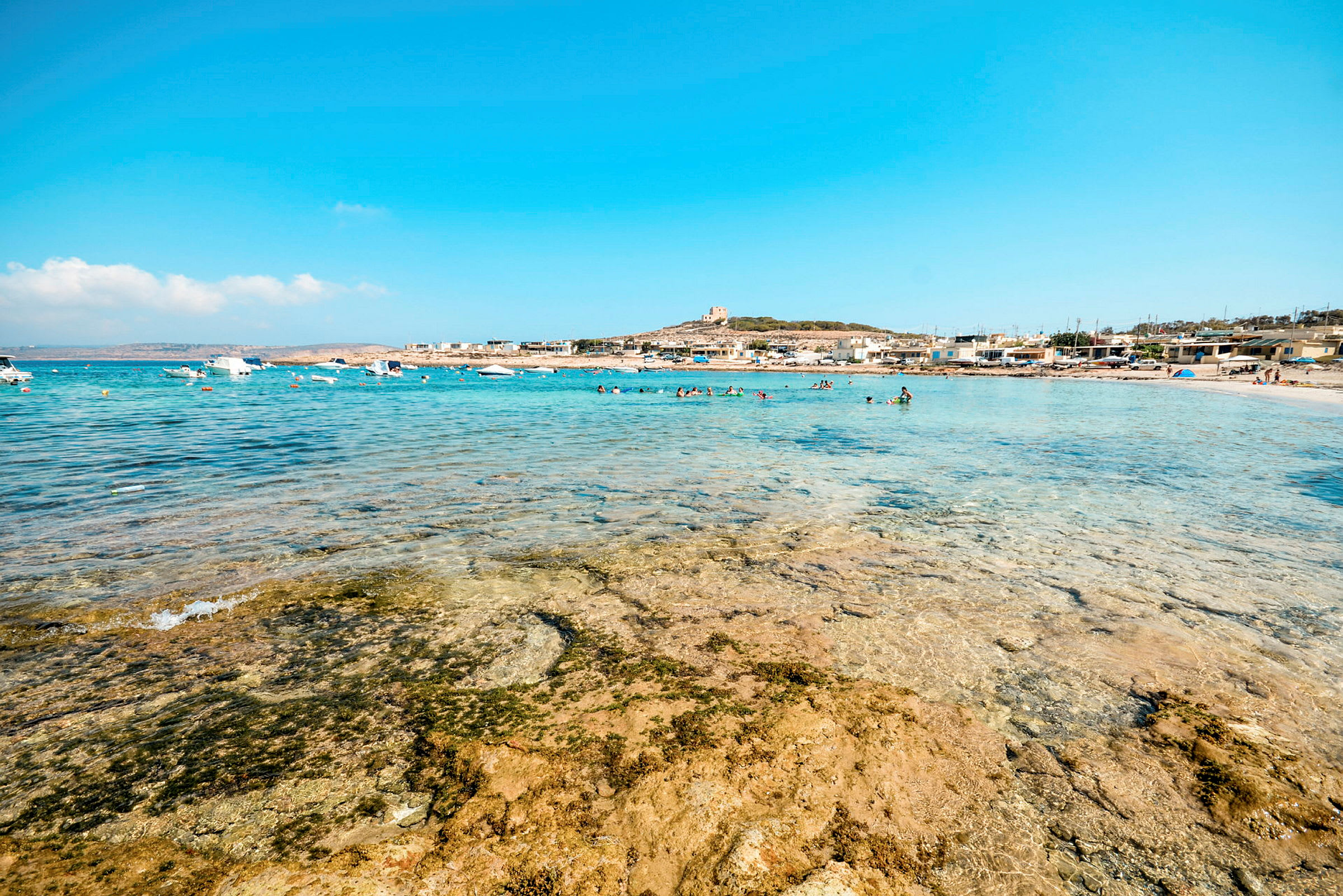 Utsikt över en strand med klart vatten och i bakgrunden båtar, badande personer och byggnader. (AI-genererad text)