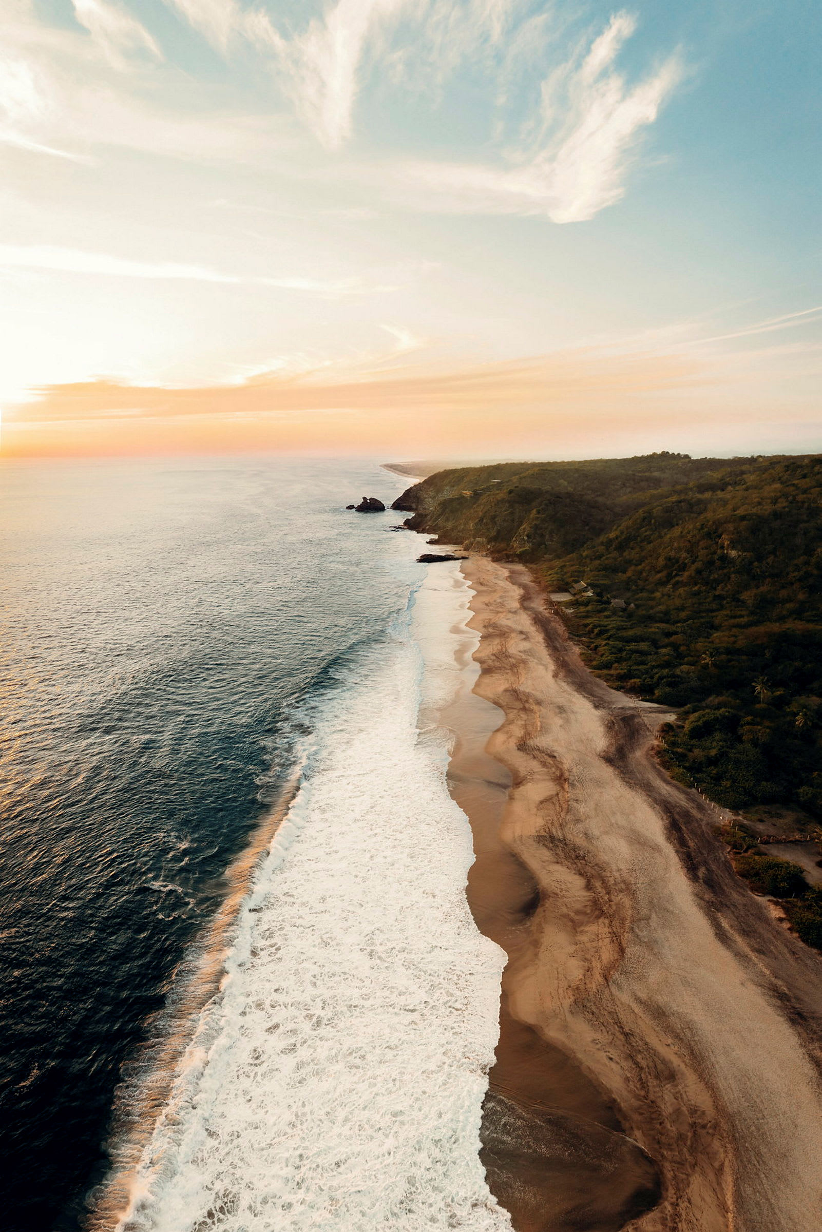 En kustlinje med en bred strand som möter vågorna från havet under en solnedgång. (AI-genererad text)