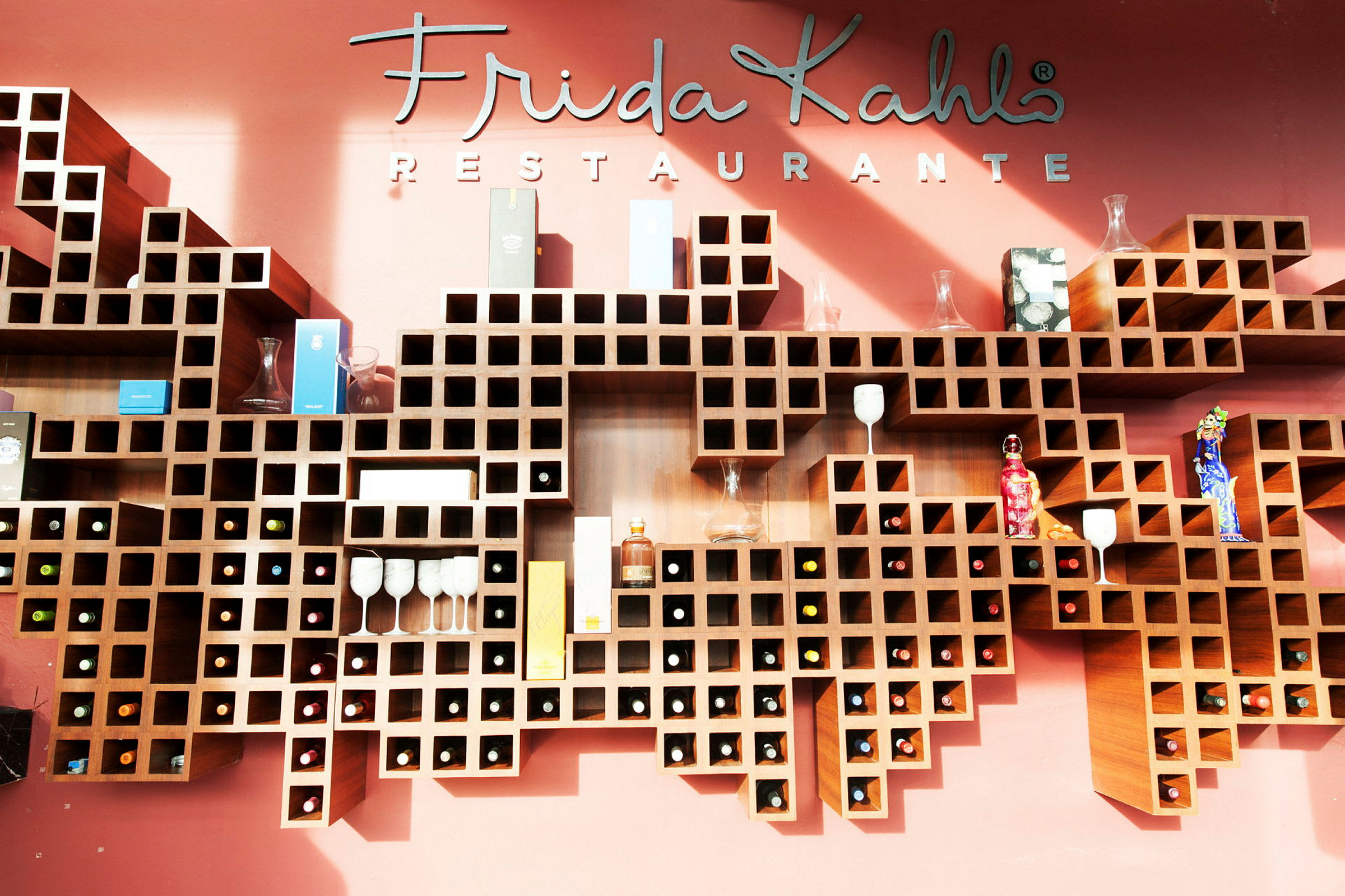 Vägghyllor med rutnätsmönster fyllda med flaskor, glas och prydnadsföremål framför en vägg med texten "Frida Kahlo Restaurante". (AI-genererad text)