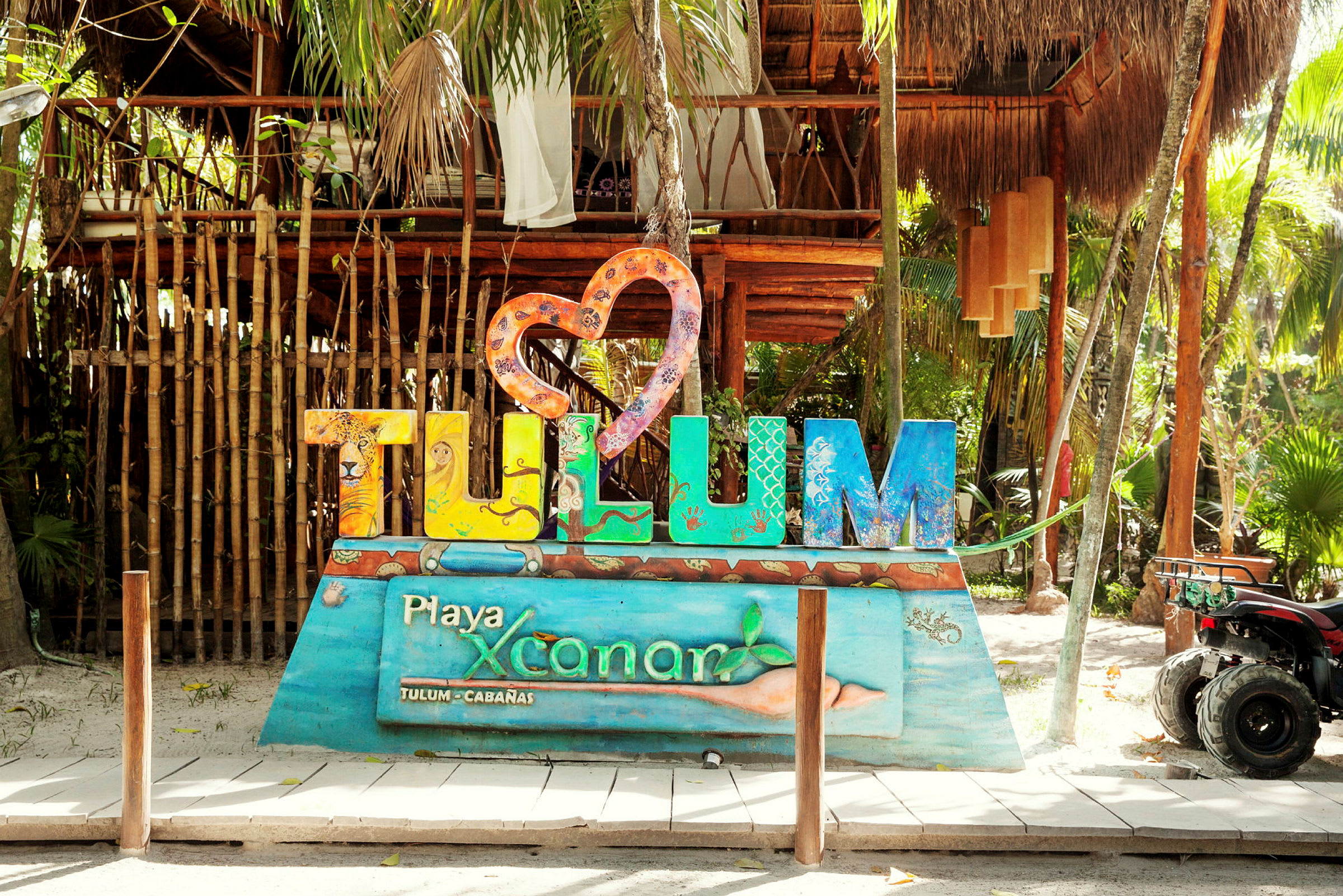 Ett färgglatt Tulum-skylt framför en tät skog och en träbyggnad. (AI-genererad text)